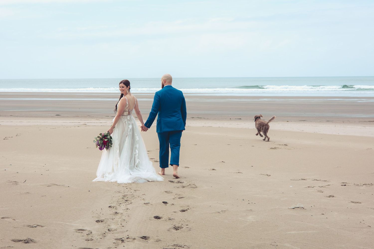 Bride and groom walk hand in hand on beach as their dog runs freely nearby.