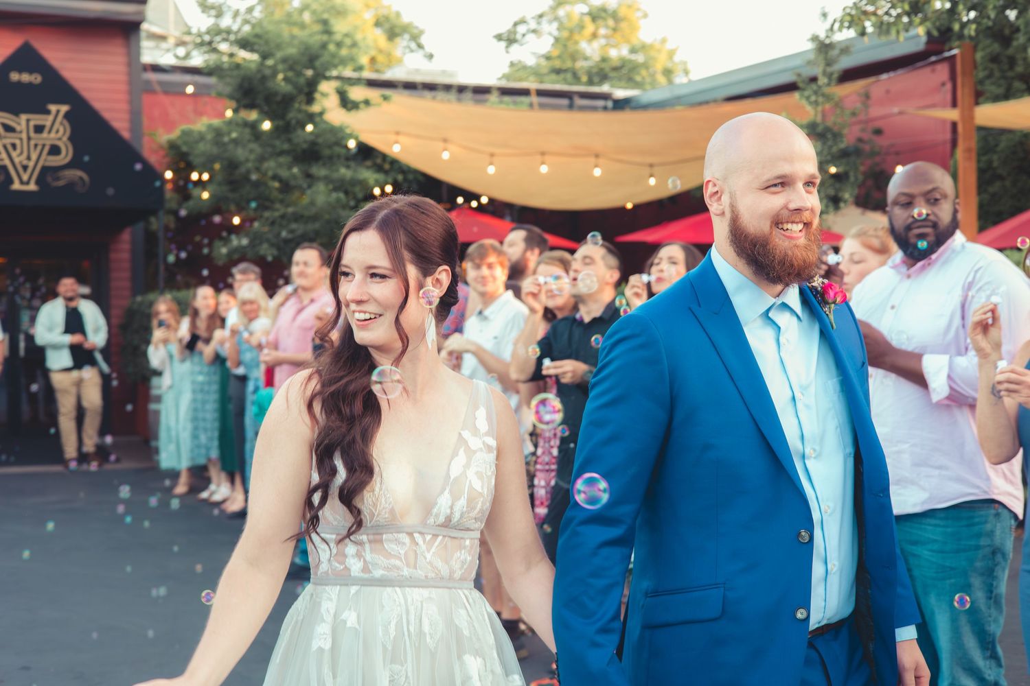 Newlywed couple walks through cheering guests with bubbles at an outdoor wedding reception at dusk.