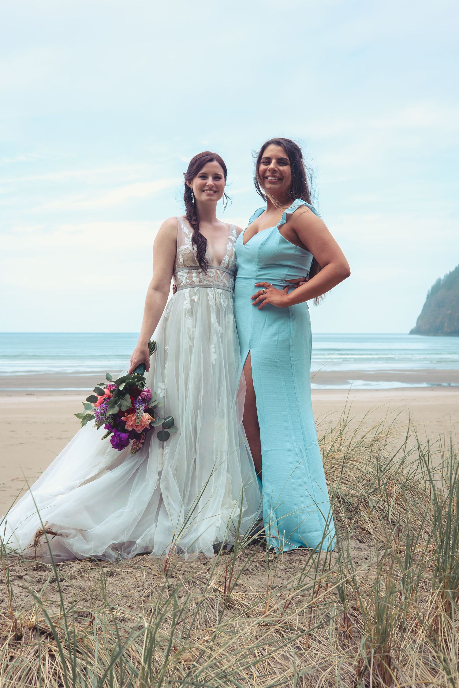 Bride holding vibrant bouquet and bridesmaid in aqua gown smiling on a beach.