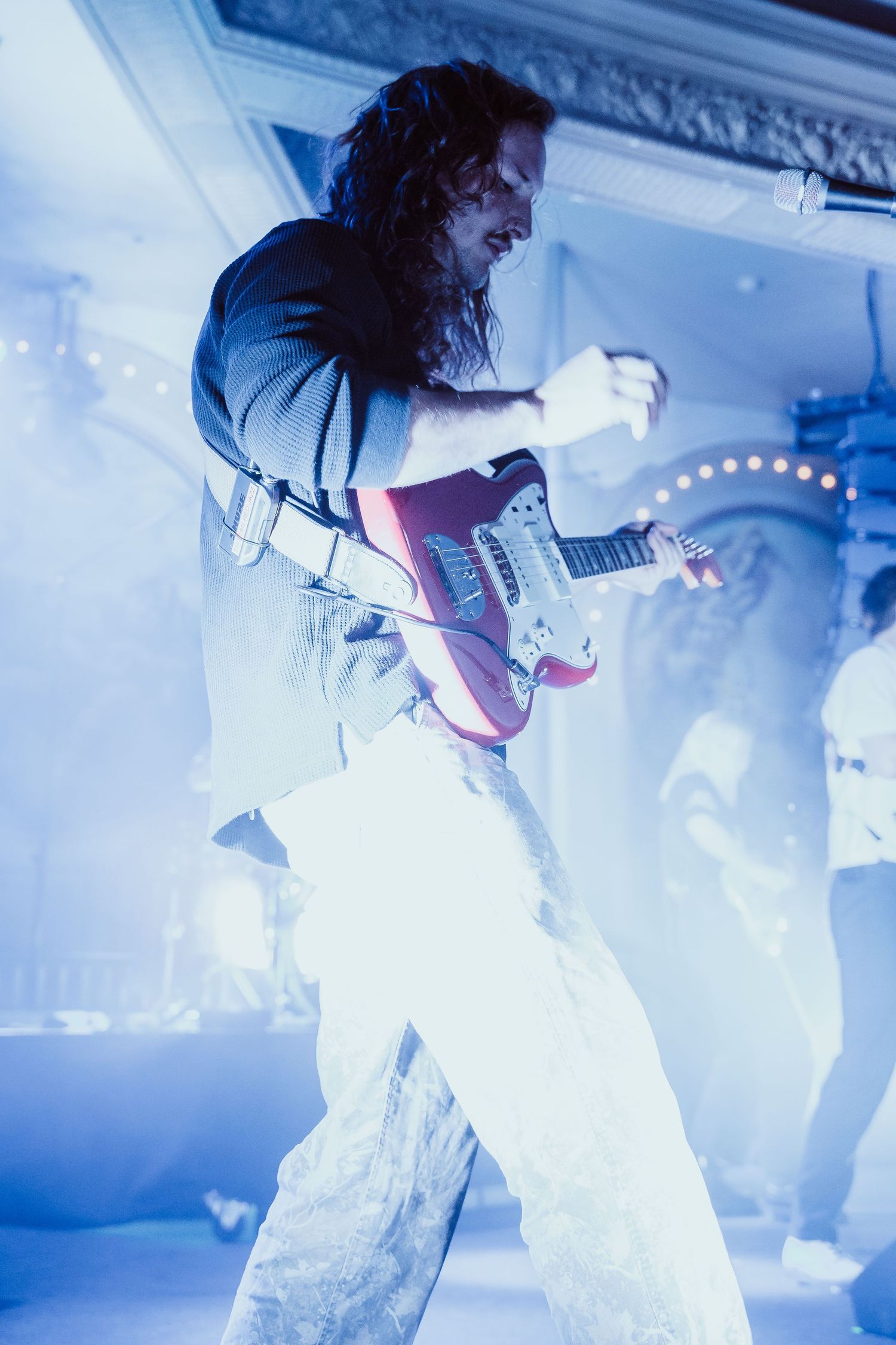 Long-haired guitarist playing red electric guitar on stage bathed in blue concert lighting.