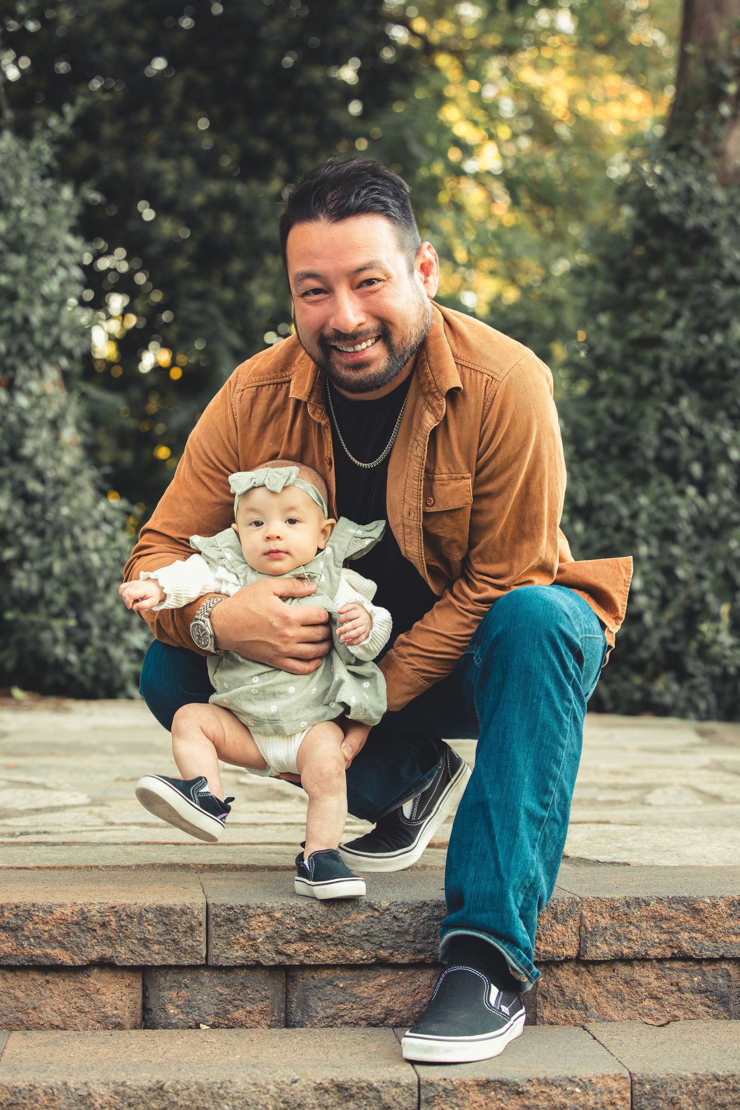 Smiling father in brown shirt sitting on stone steps outdoors with his baby daughter in green dress.
