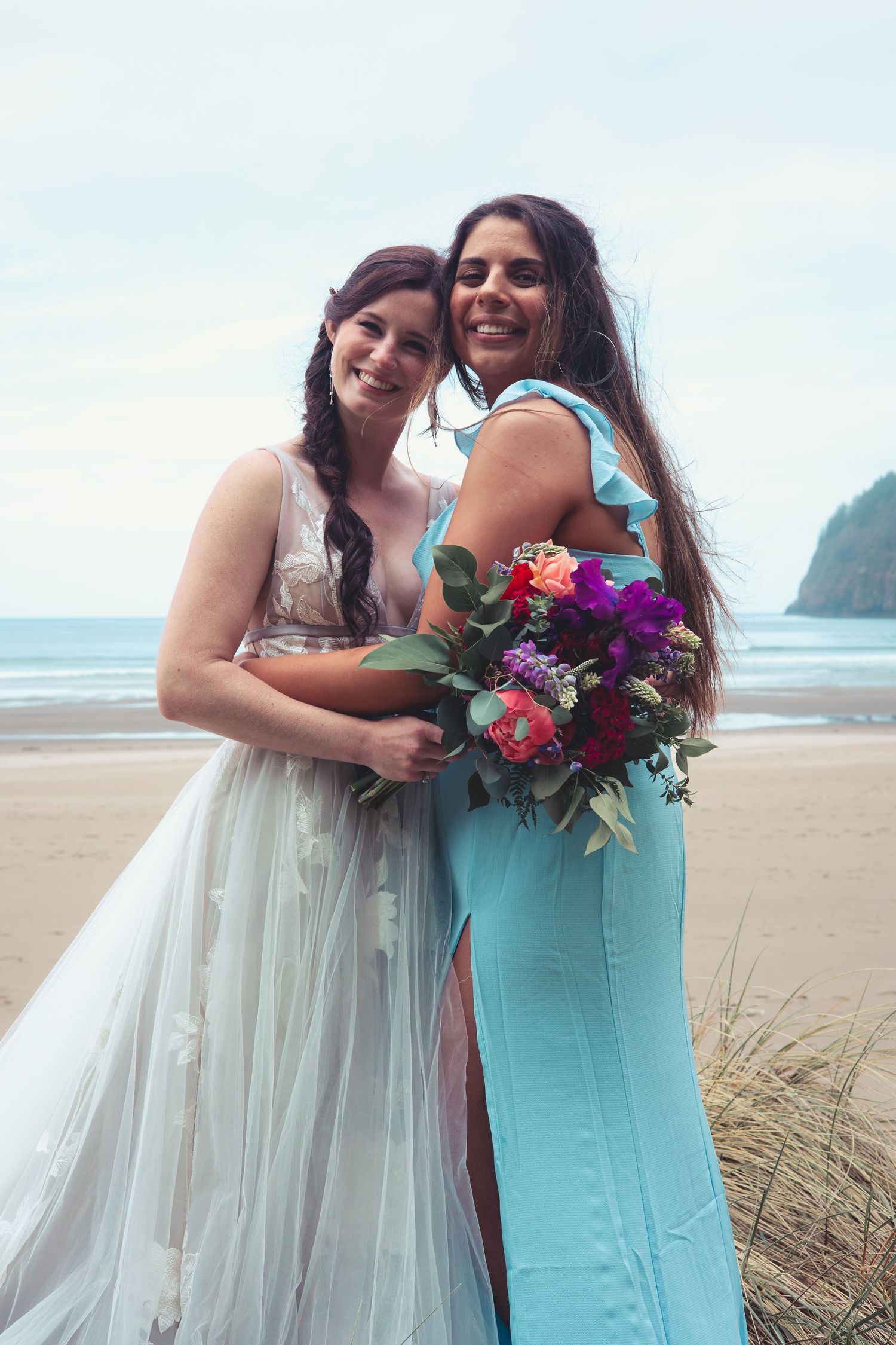 Bride and bridesmaid in blue dress hugging while holding colorful bouquet on beach.