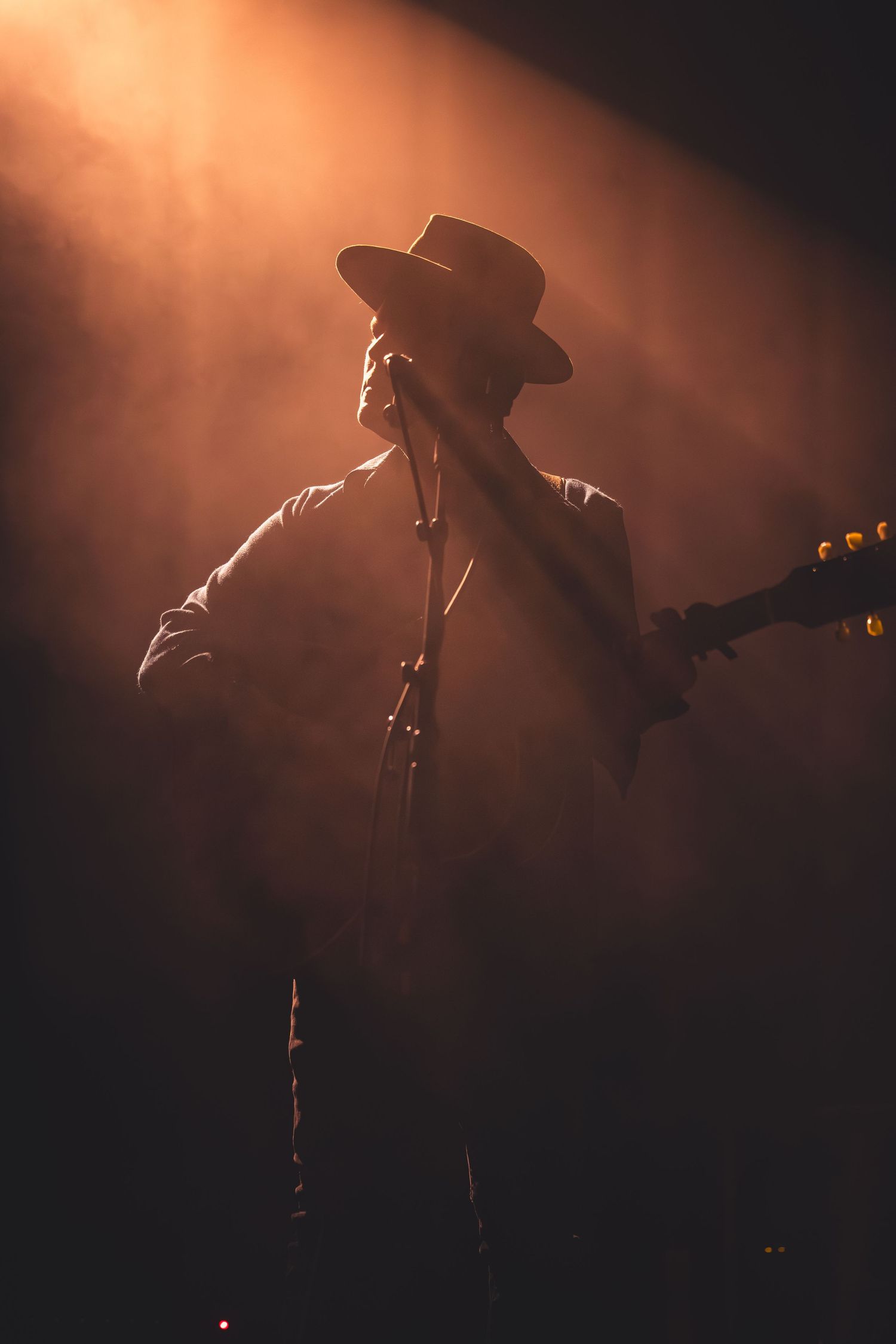 Silhouetted guitarist in cowboy hat performing on stage under warm amber spotlight with dramatic smoke effects.