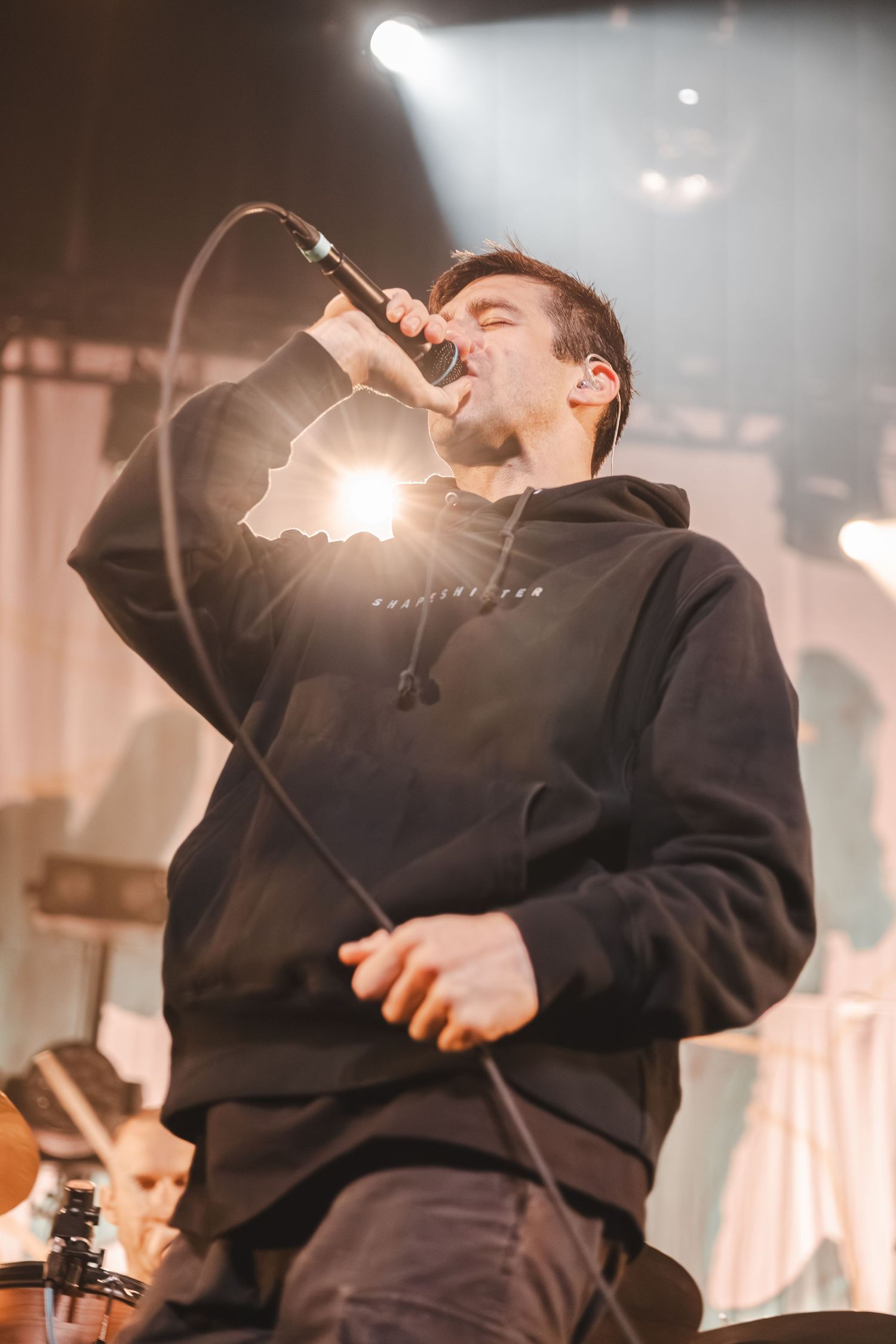 Young male vocalist singing passionately into a handheld mic on a brightly lit concert stage.