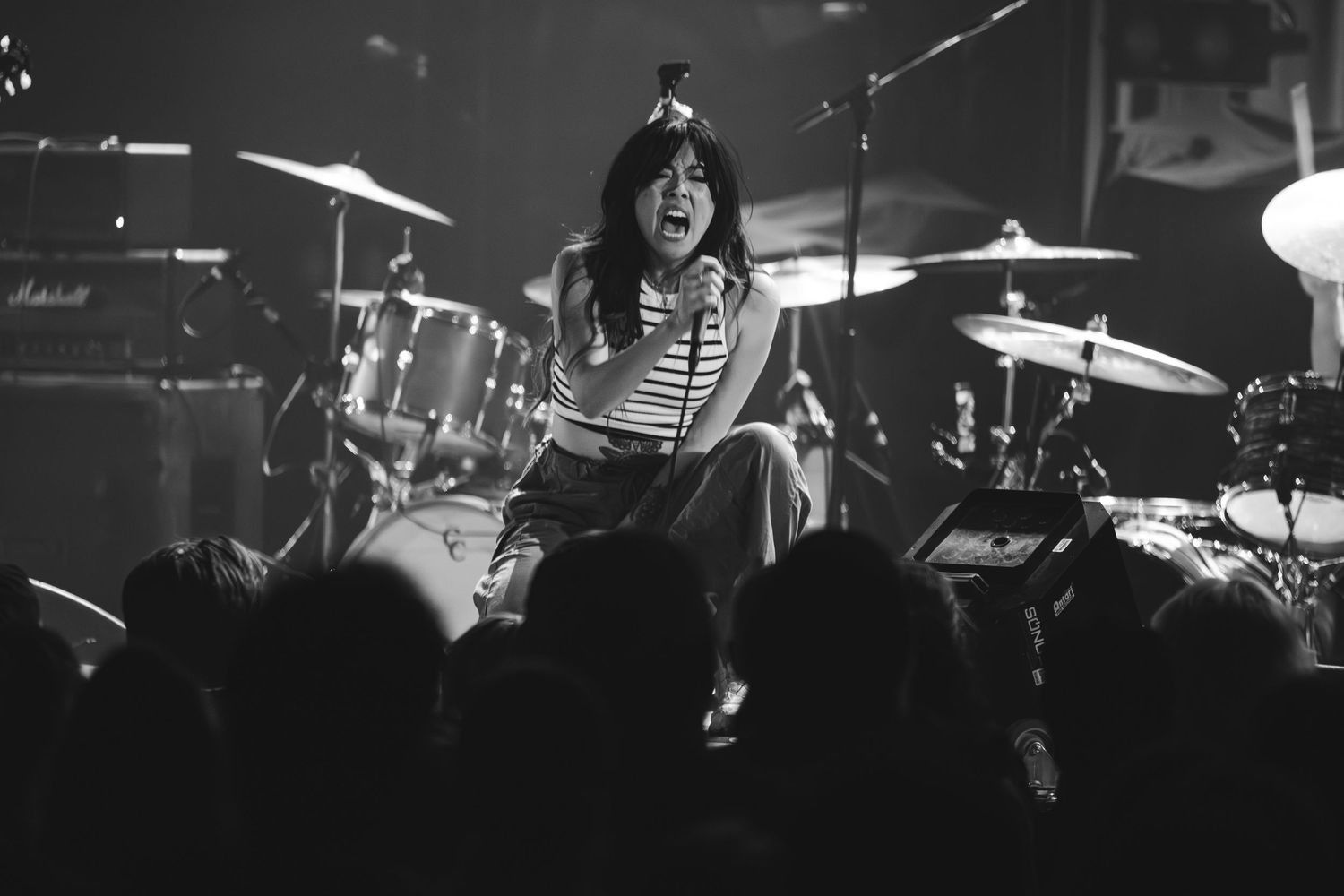 Black and white photo of female vocalist singing passionately on stage with drum kit visible behind her.