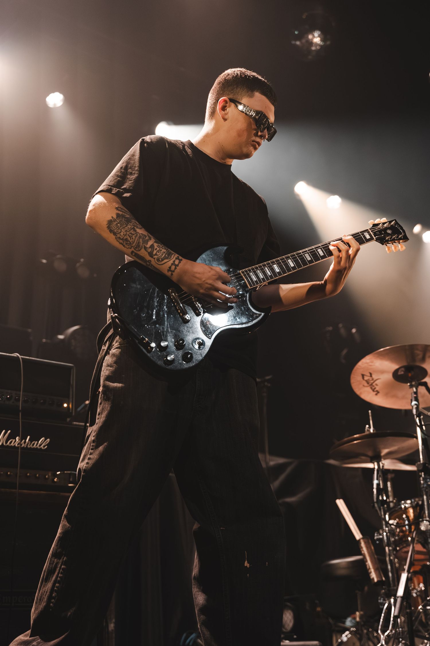 Tattooed male guitarist with sunglasses plays blue electric guitar on stage near Marshall amplifier.