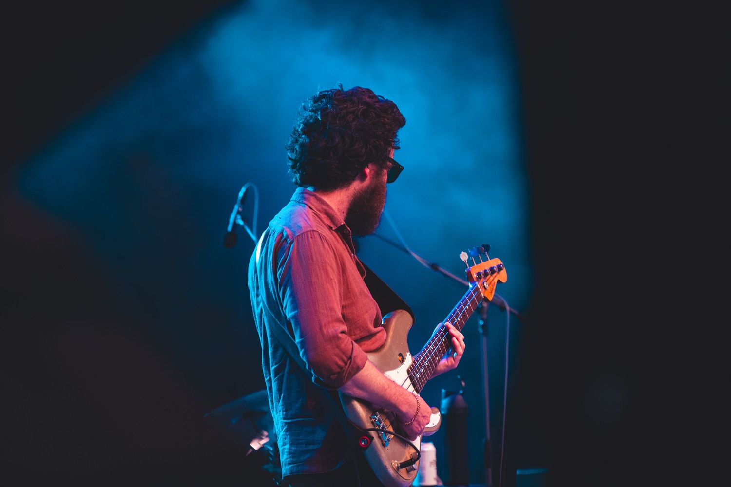 Curly-haired bearded bassist performing on a dramatic blue-lit smoky concert stage.