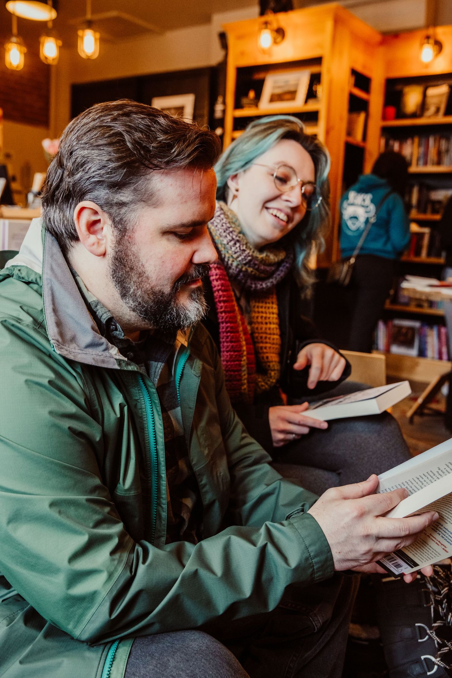 A bearded man reads a book as a smiling blue-haired woman looks on in a cozy bookstore.