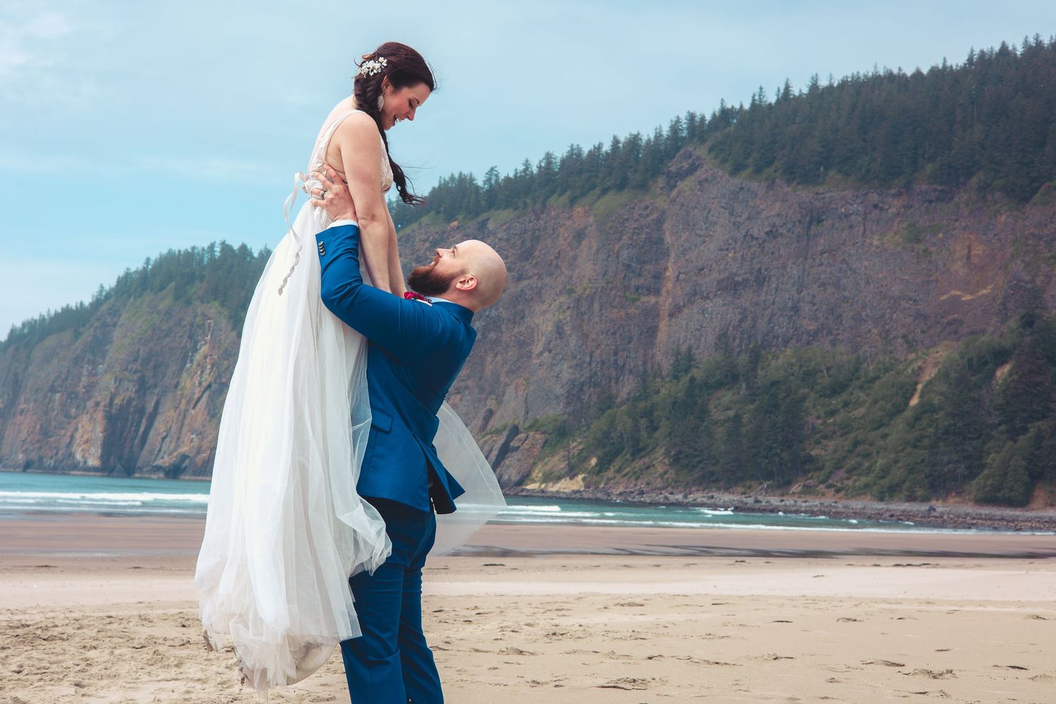 Groom in blue suit lifts smiling bride in white gown high on sandy beach with forested cliffs behind.
