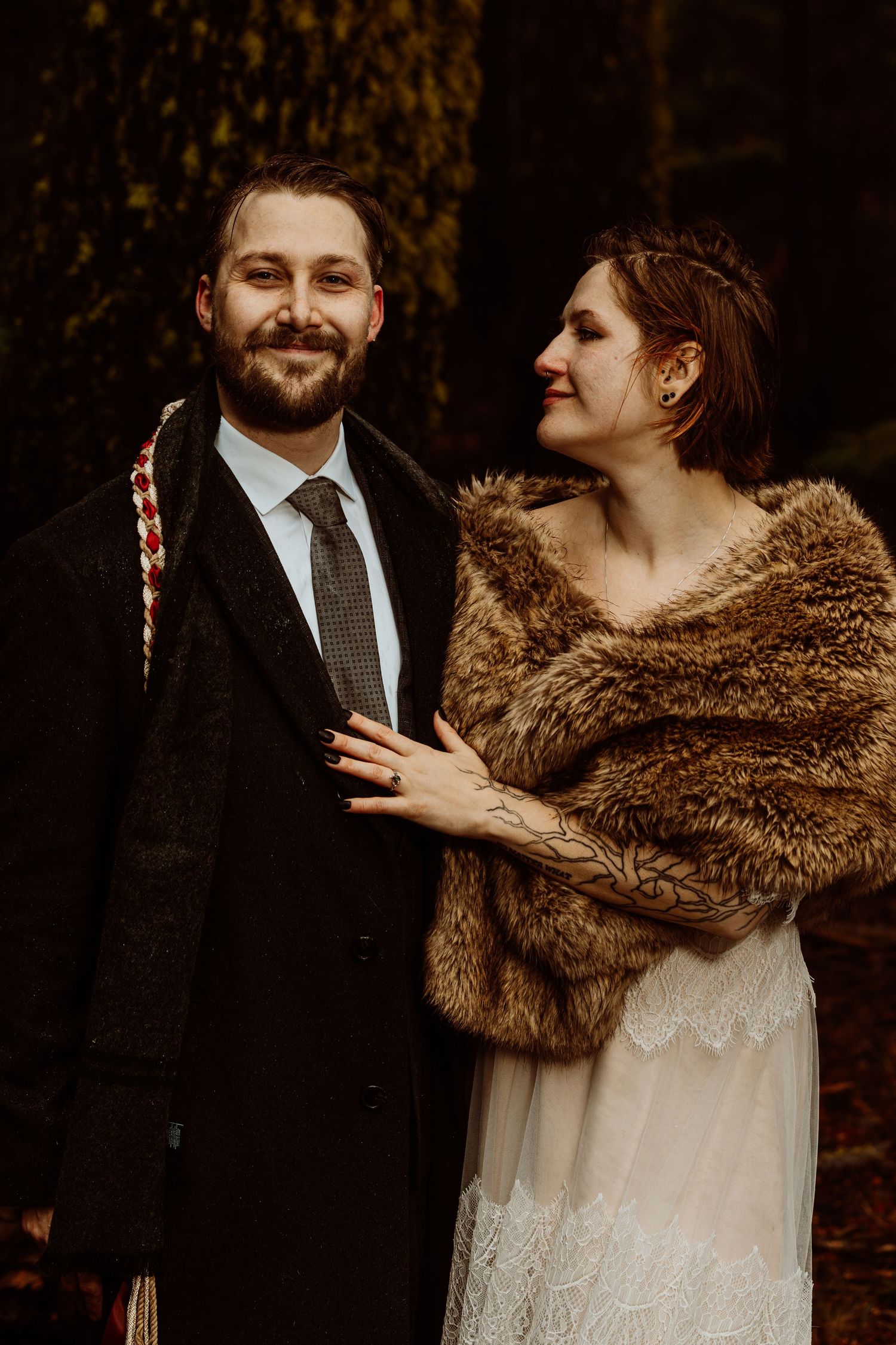 Wedding couple posing in forest, bride in white lace dress with fur wrap, groom in dark suit with decorative sash.