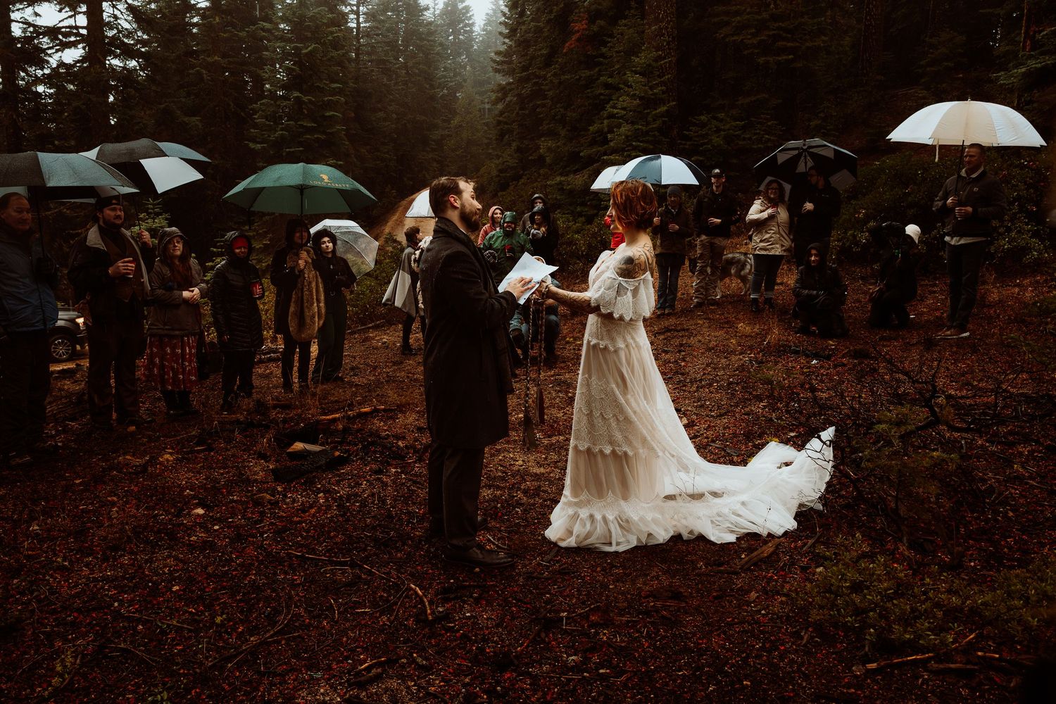 Outdoor forest wedding ceremony in the rain with guests holding umbrellas surrounding the couple.