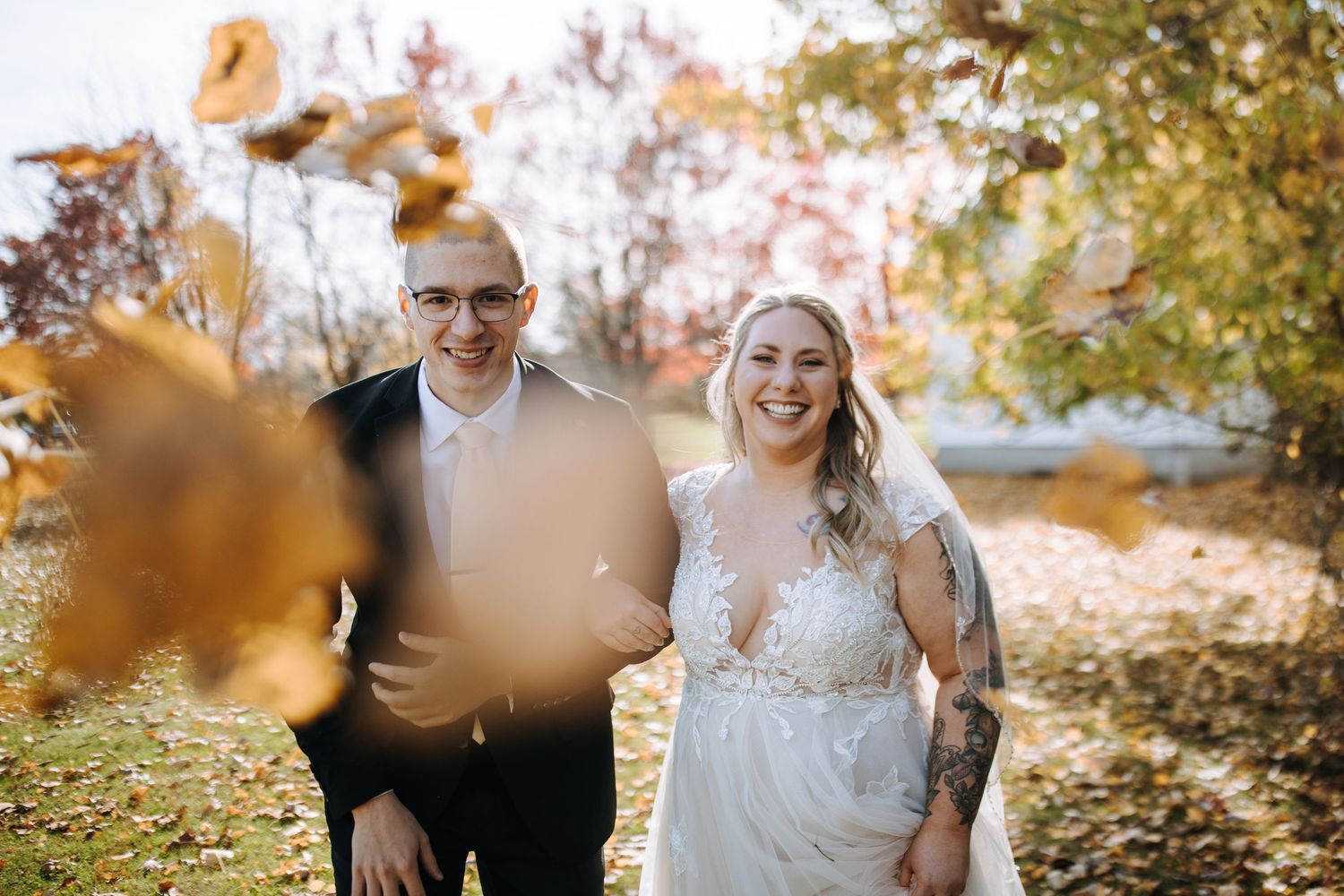 A wedding couple laughs together surrounded by golden autumn leaves in a natural outdoor setting.