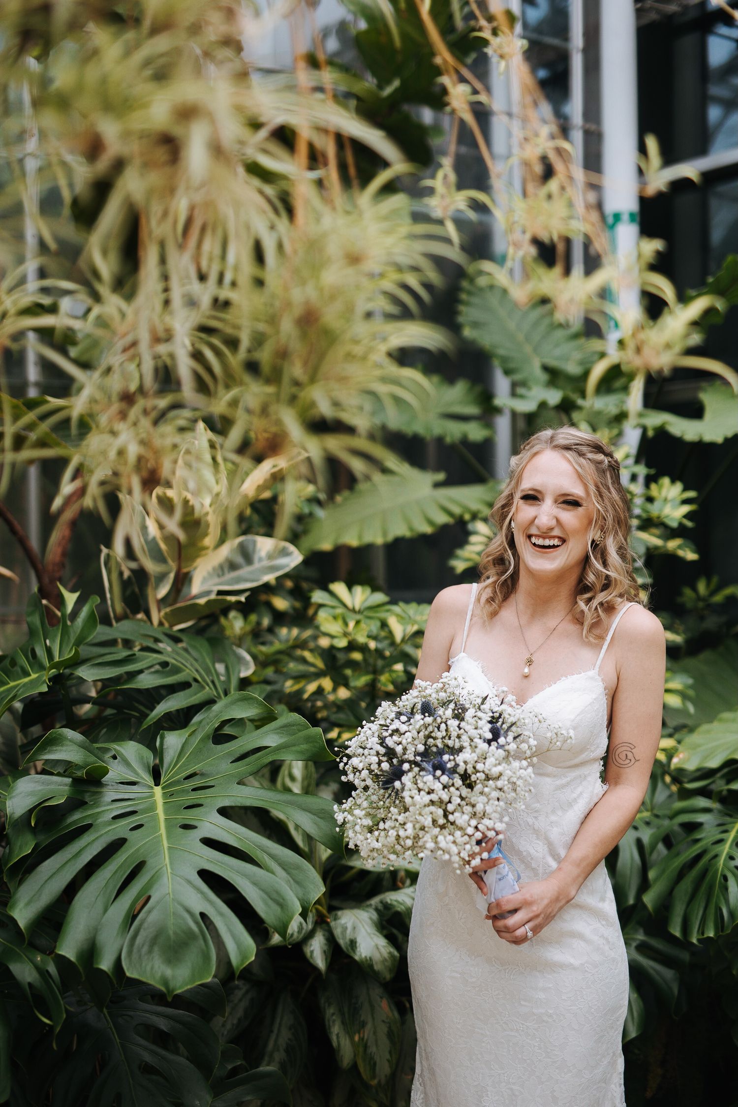A white wedding dress is showcased among tropical monstera leaves in a greenhouse setting.