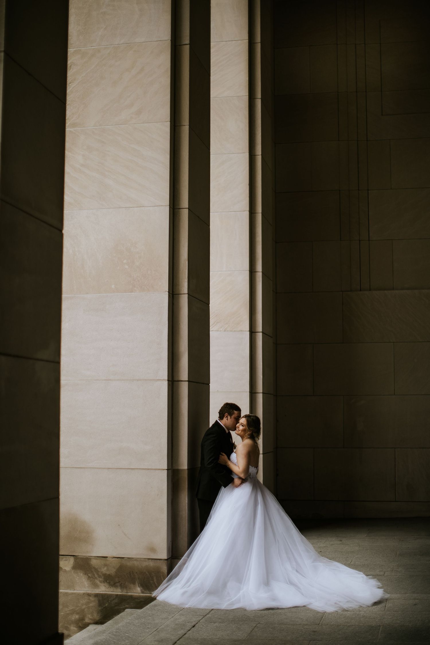 Dramatic architectural portrait of a bride in a flowing white gown against stone columns.