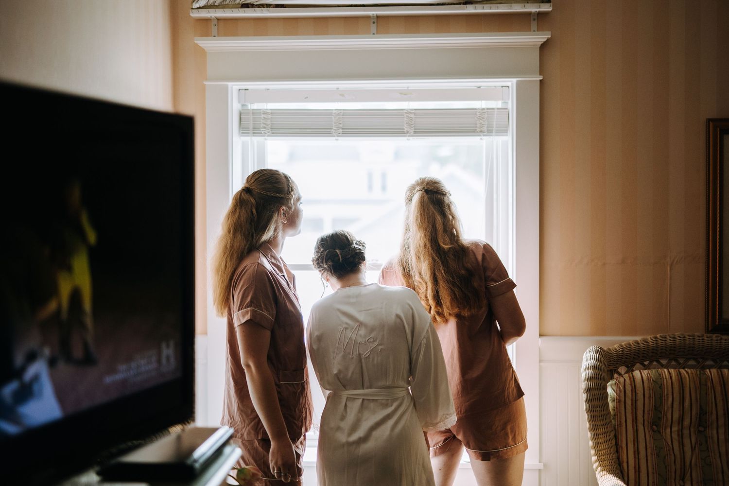 Three women look out a bright window during wedding preparations.