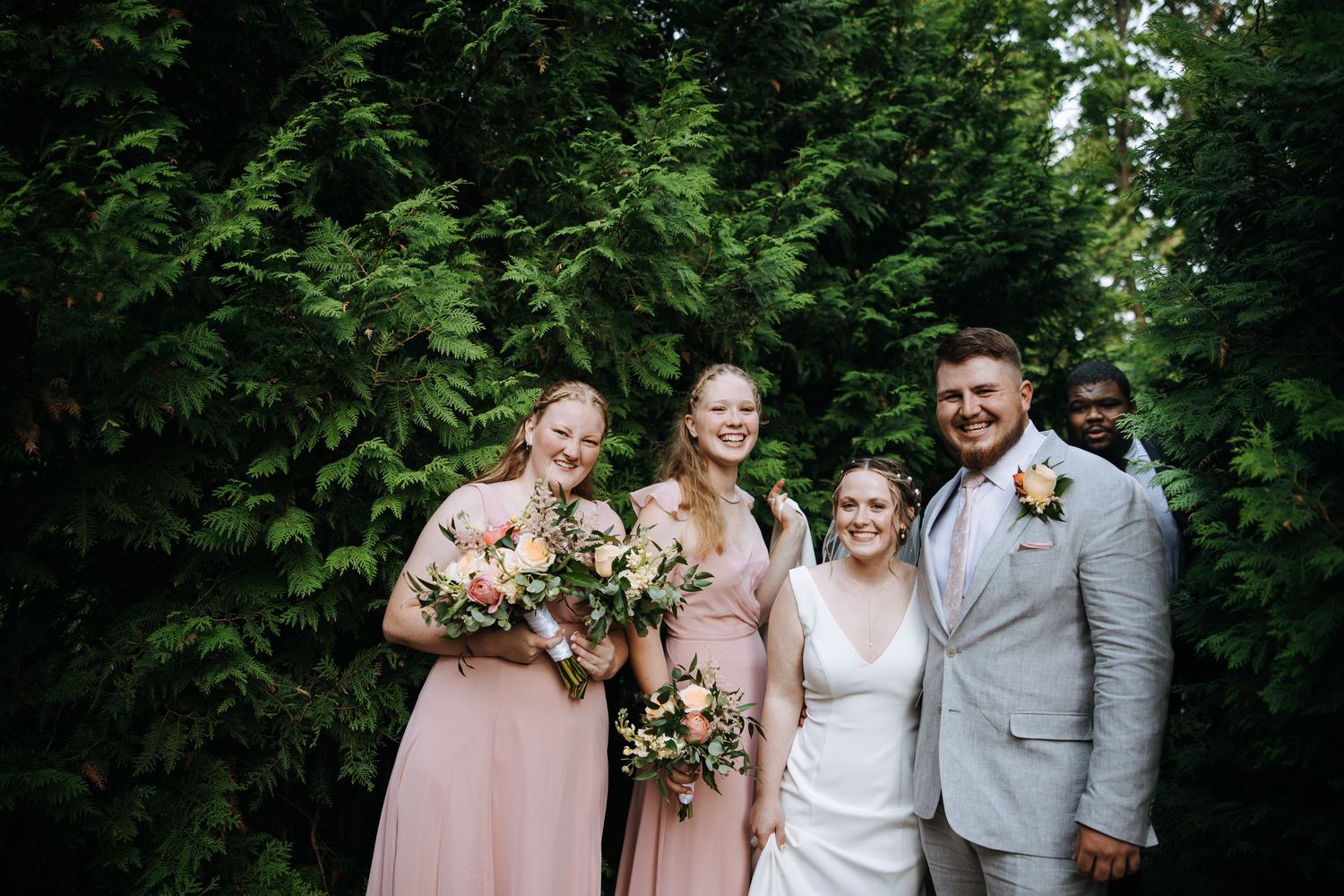 Wedding party poses together on garden path surrounded by evergreen trees.