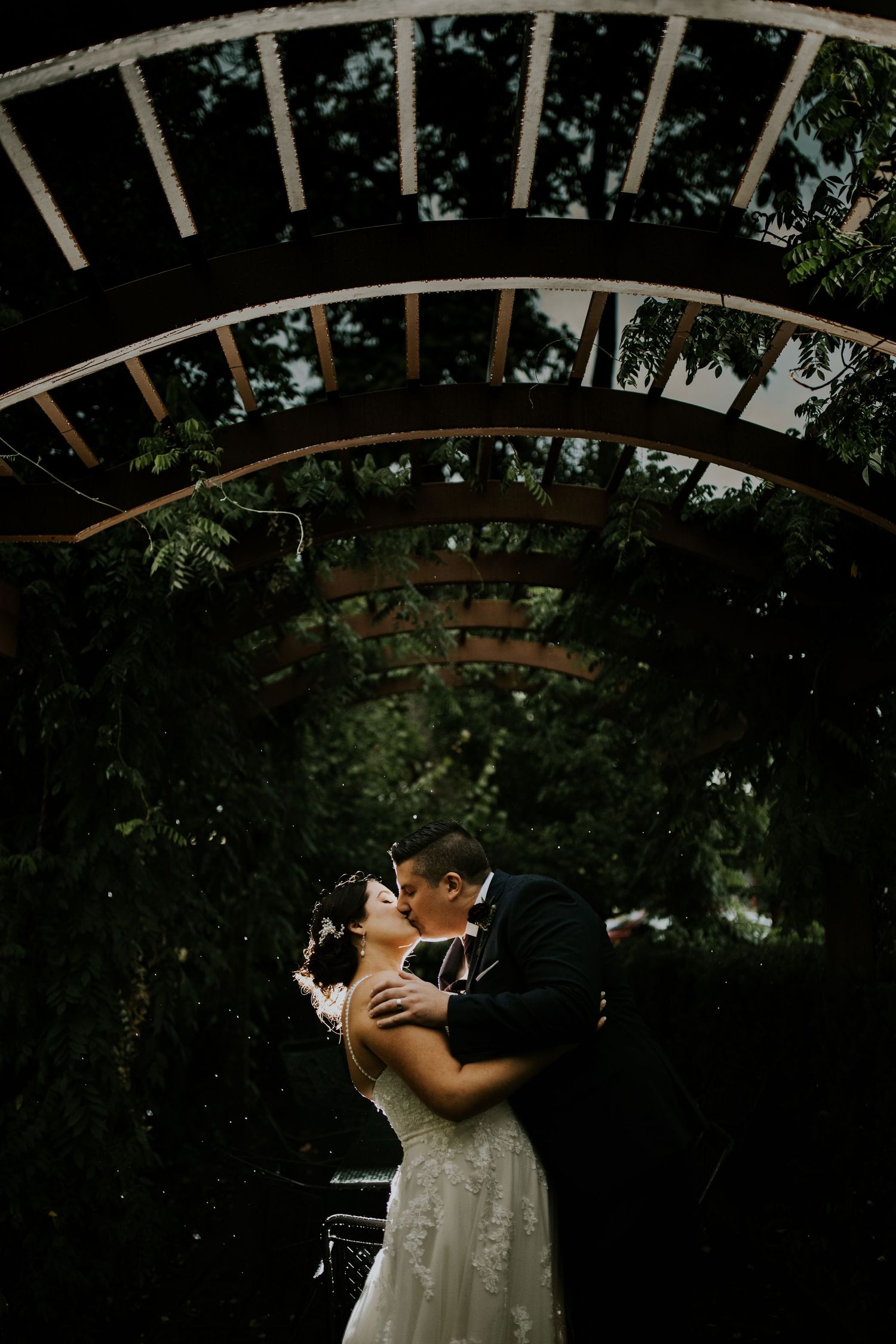 A romantic wedding kiss under a wooden garden arch surrounded by lush greenery at dusk.