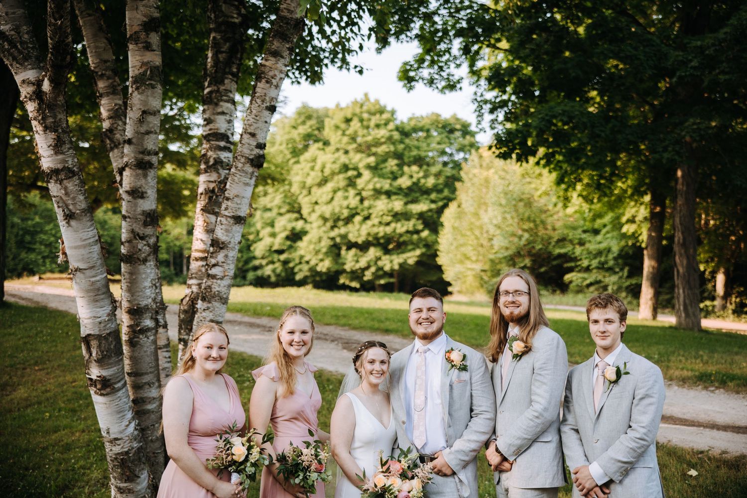 A wedding party in pink dresses and gray suits poses beneath birch trees.