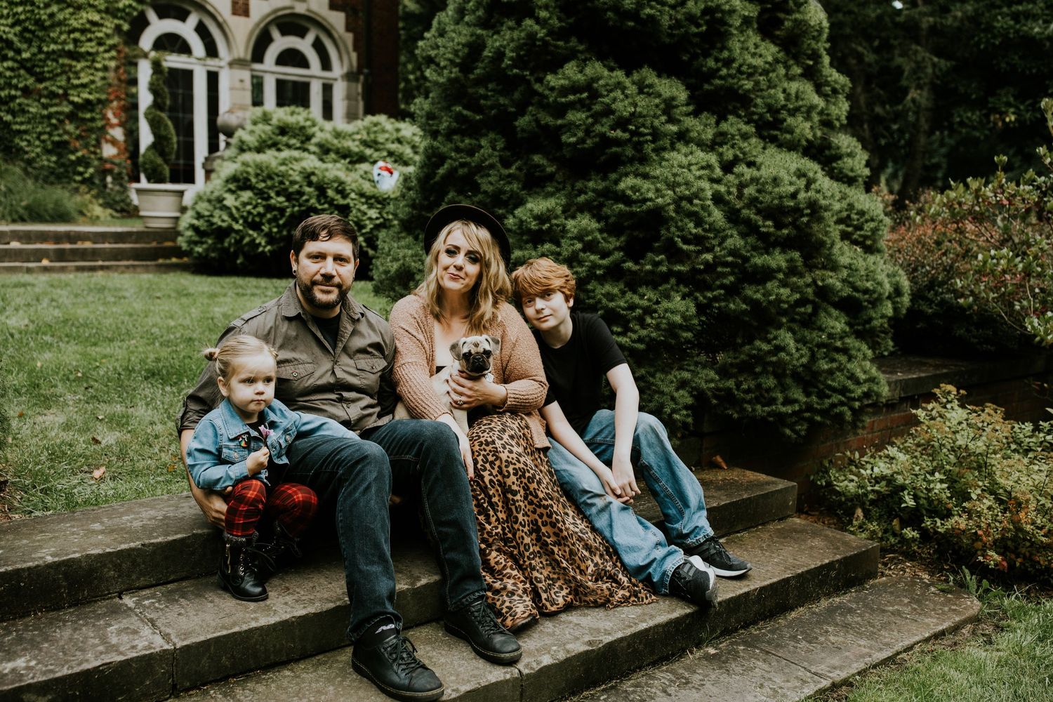 Family members sit casually on outdoor steps surrounded by green foliage.