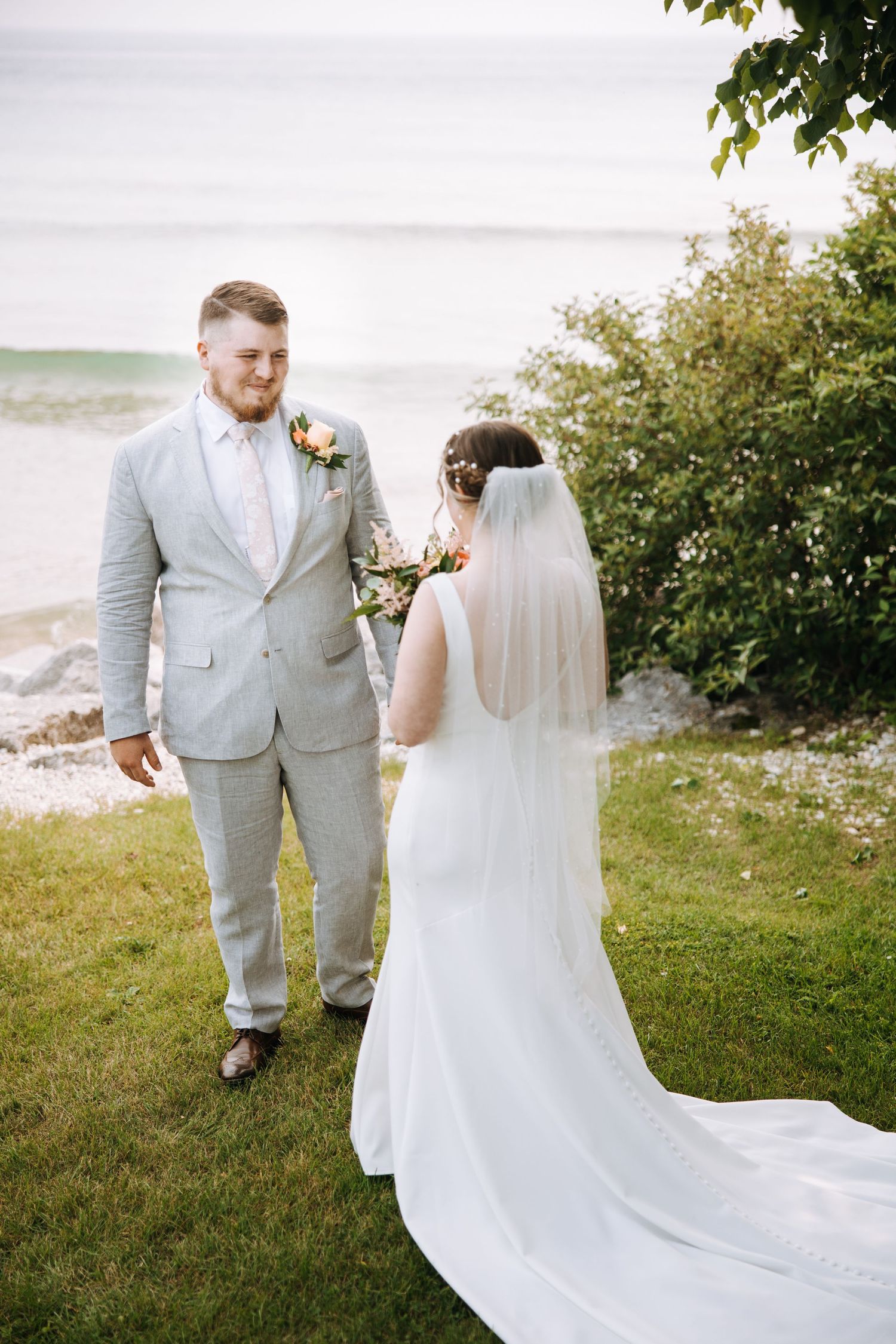 A couple in wedding attire shares a moment together on a grassy hillside overlooking the water.