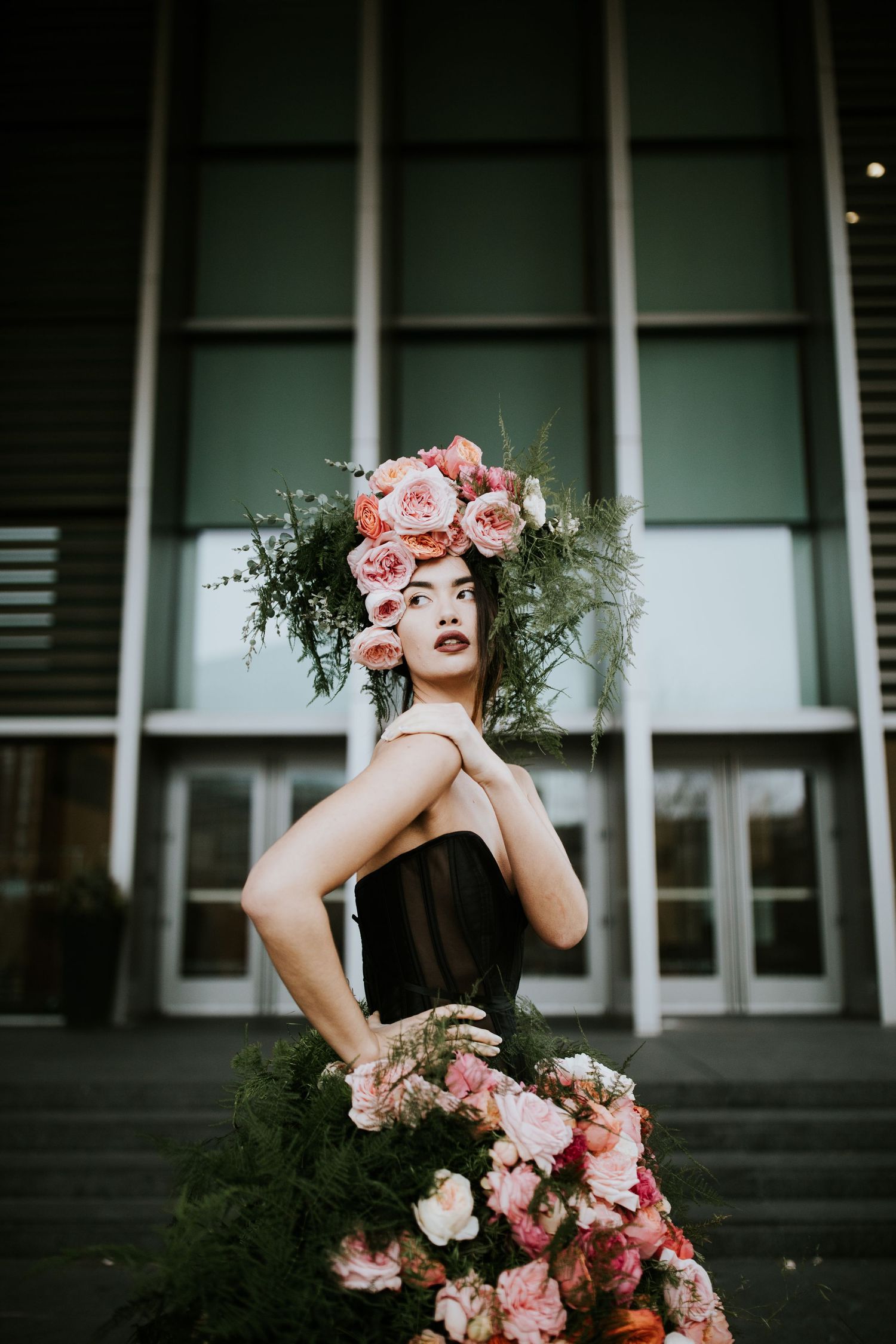 Dramatic outdoor fashion shoot with floral crown and pink roses against modern architecture.