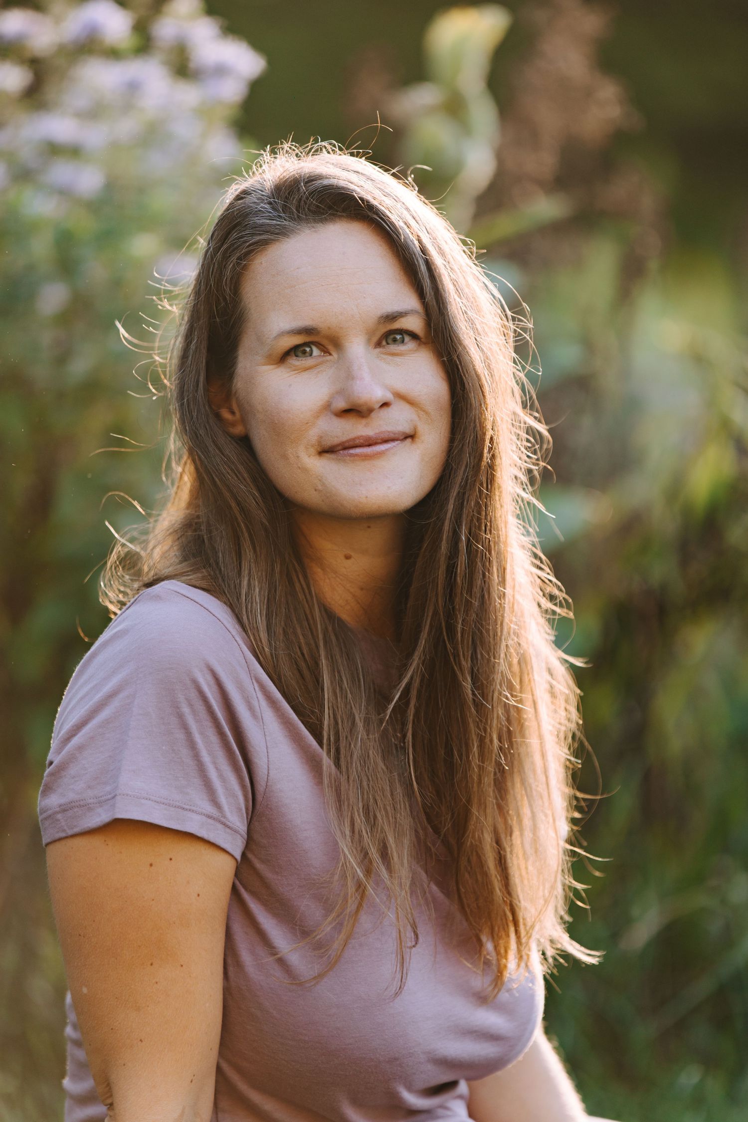 A person with long brown hair wearing a mauve t-shirt smiles while posing outdoors in natural lighting.