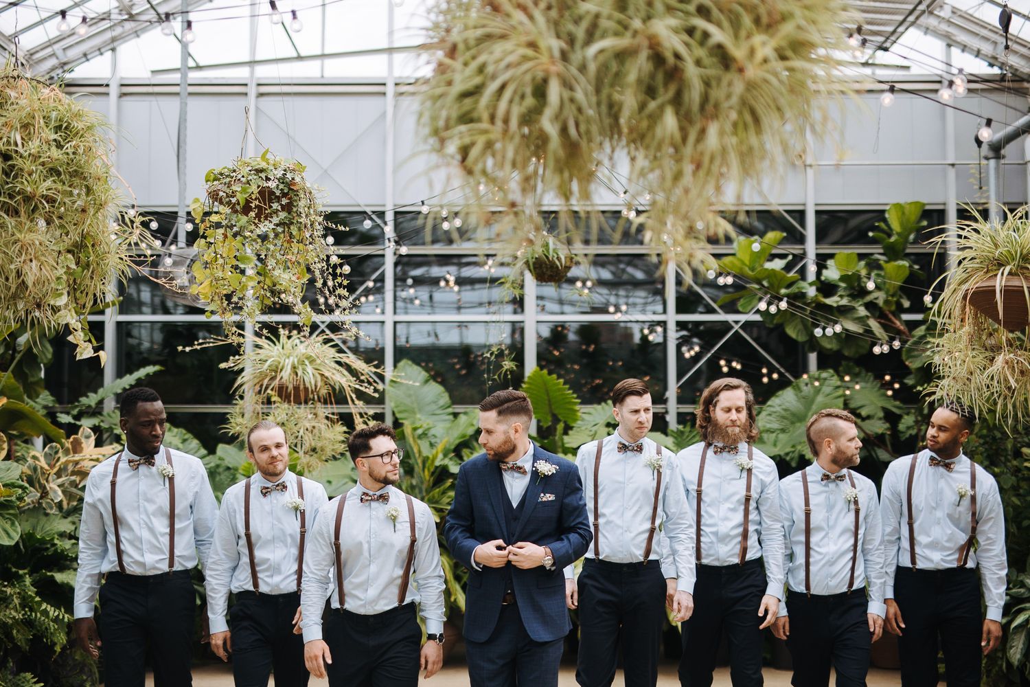 Wedding party members in suspenders and bow ties pose in front of a greenhouse backdrop.