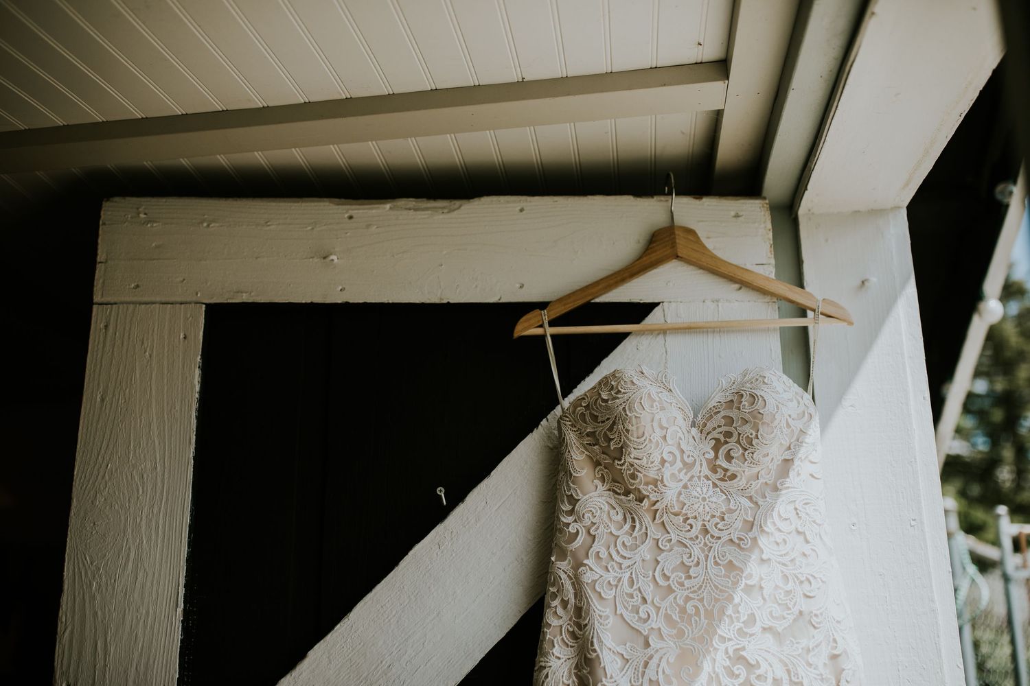 A wedding dress hangs from a wooden hanger against a white wall, showcasing its delicate lace and elegant design.