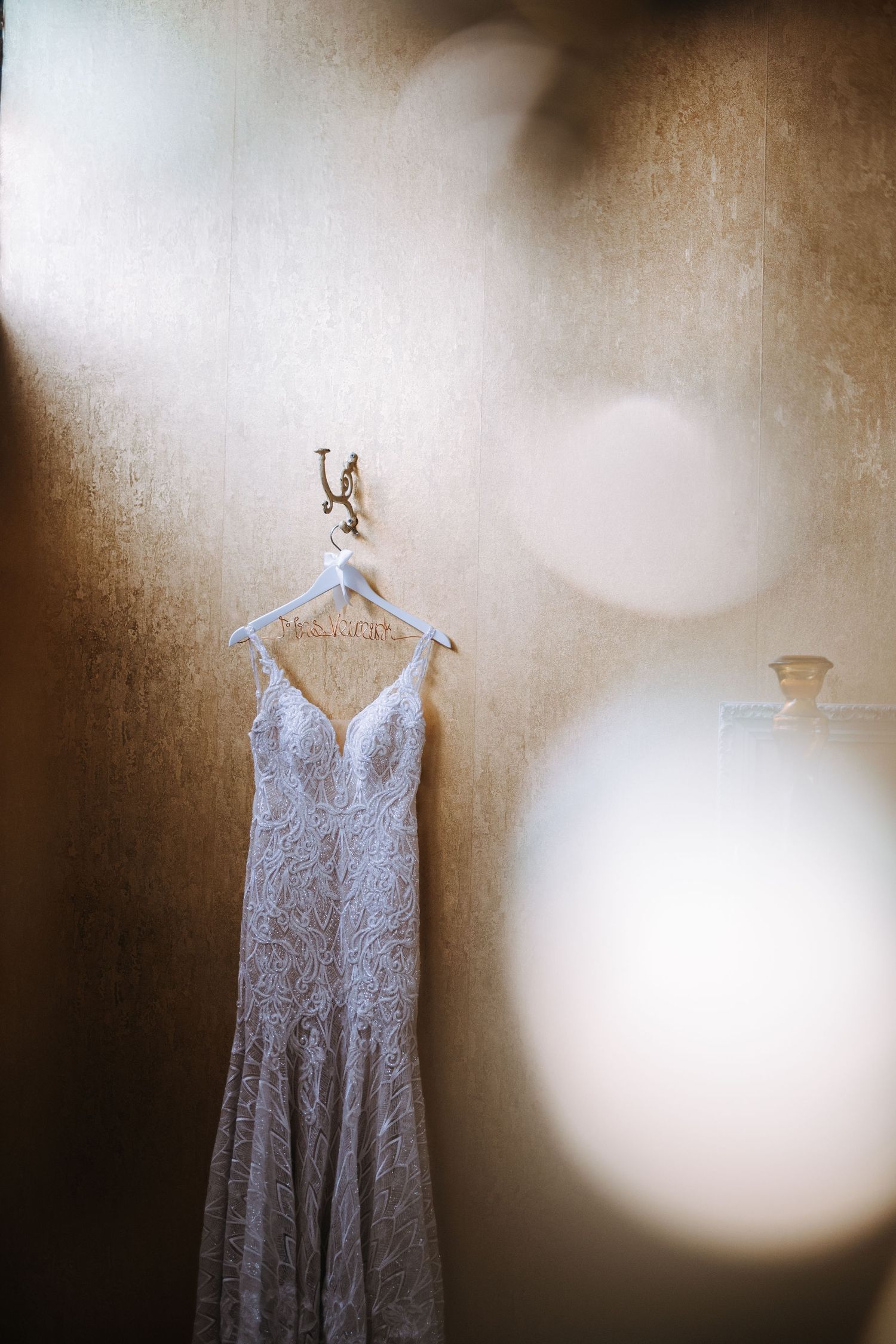 A white lace wedding dress hangs on a decorative hanger against a textured light background.