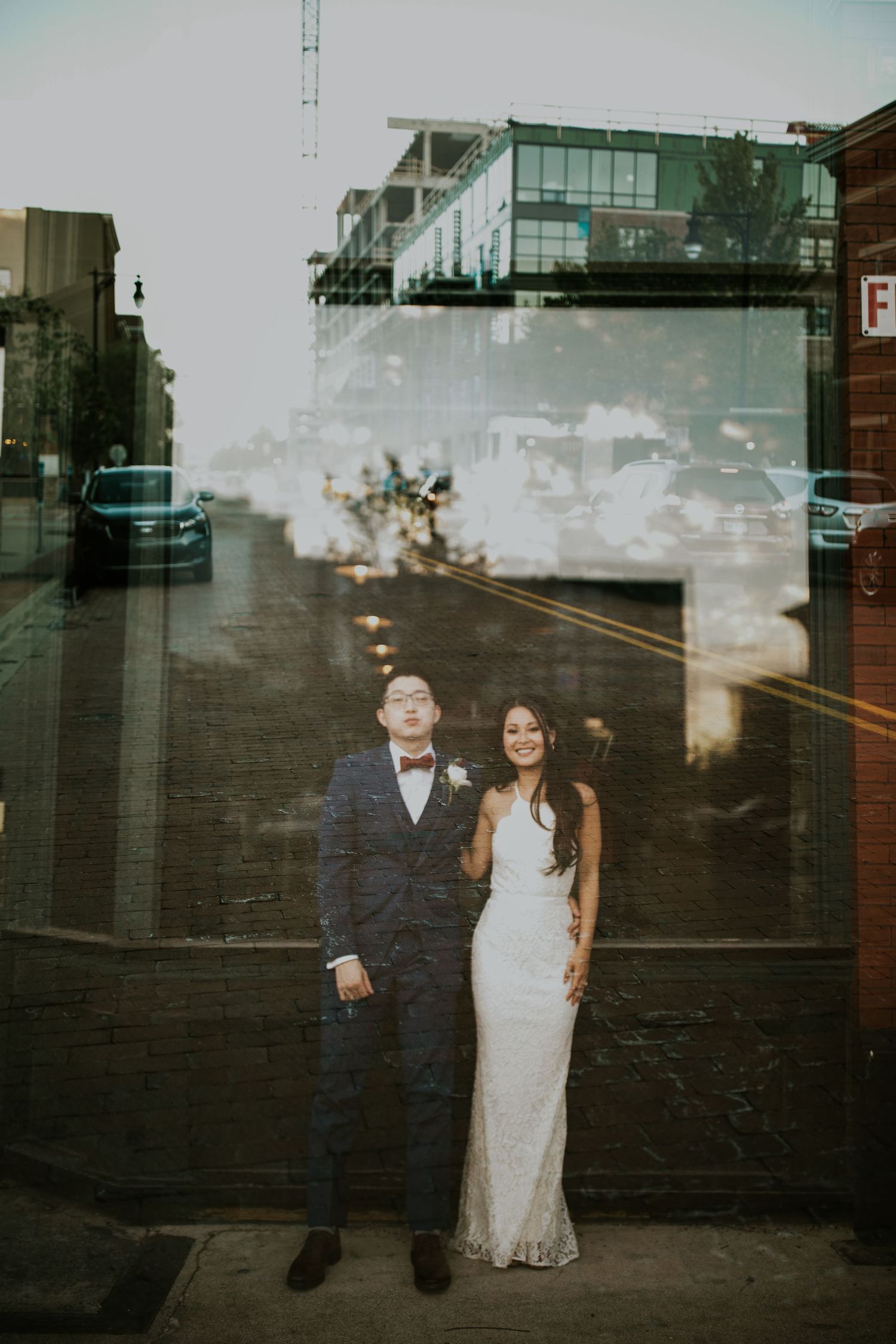 A couple in wedding attire poses against a dark brick wall in a moody urban setting.