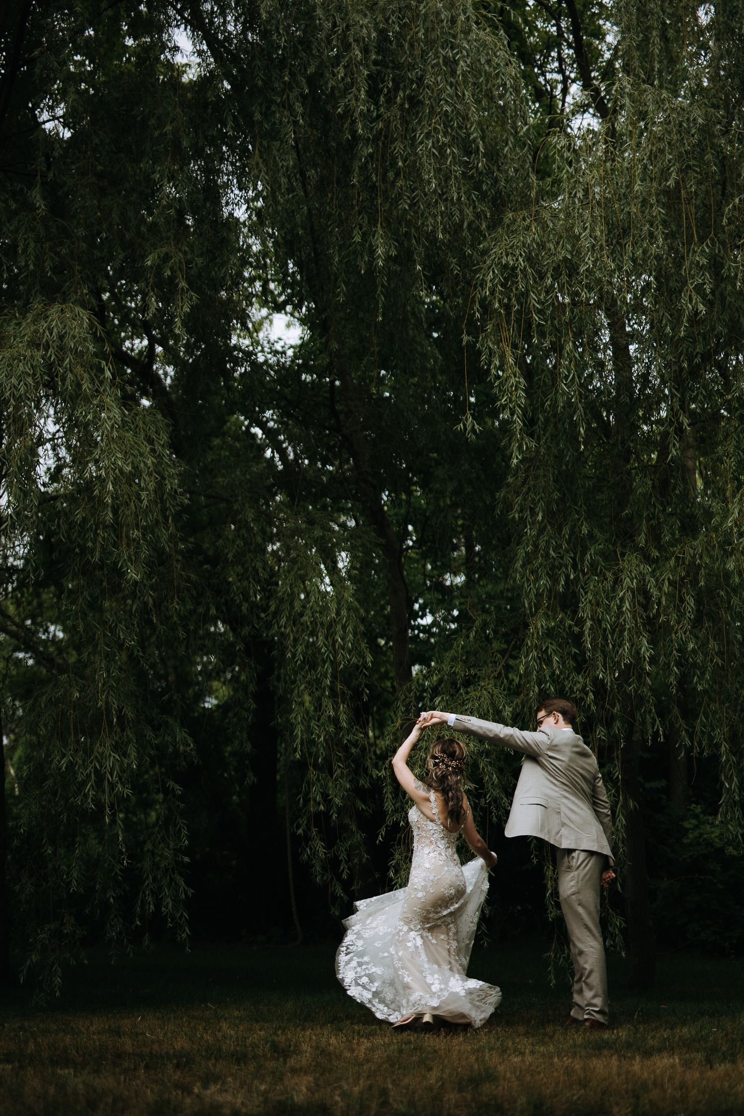 Dancing couple twirling beneath weeping willow trees at outdoor reception.