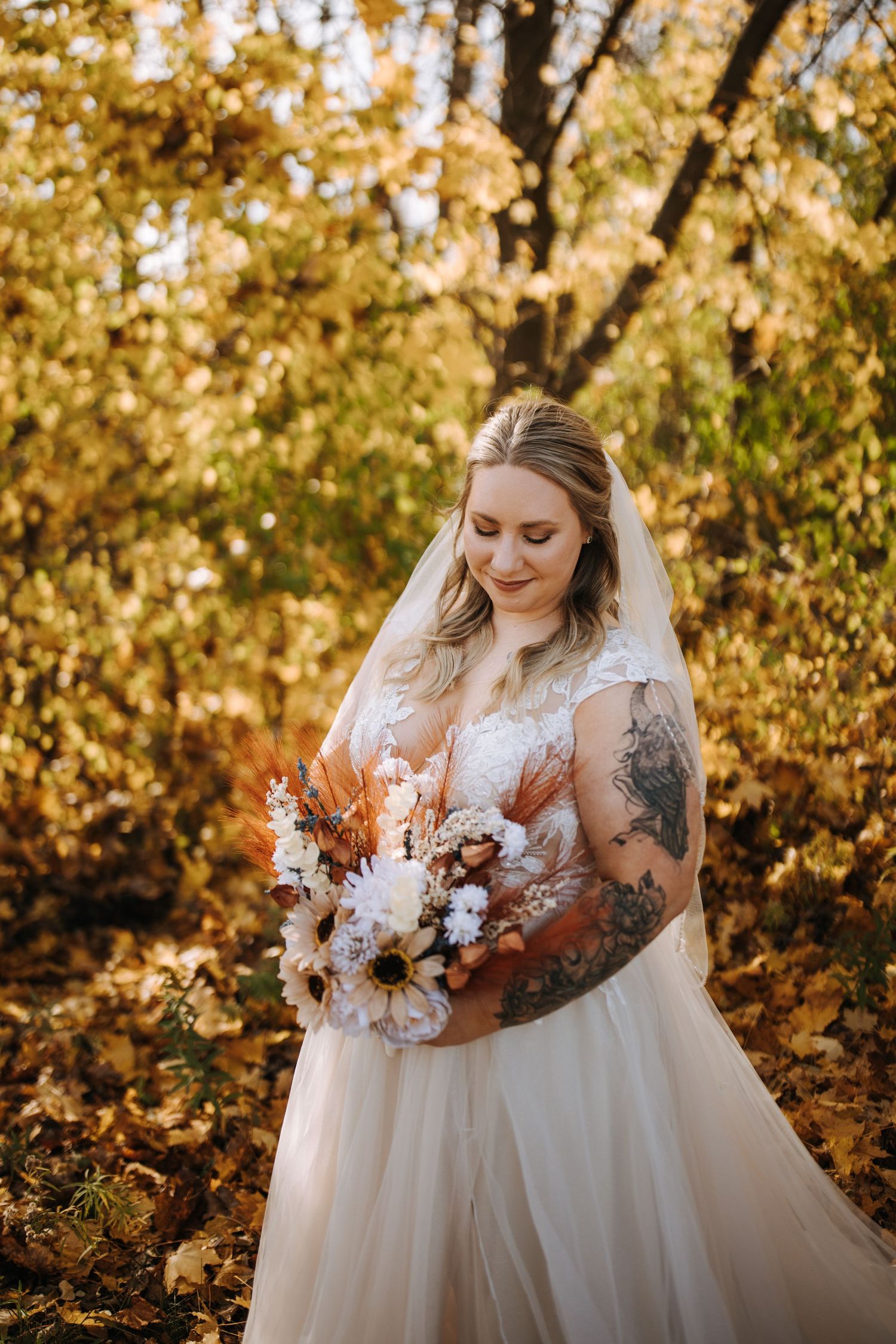 A bride in a white lace gown holds an autumn-themed bouquet against a backdrop of golden fall foliage.