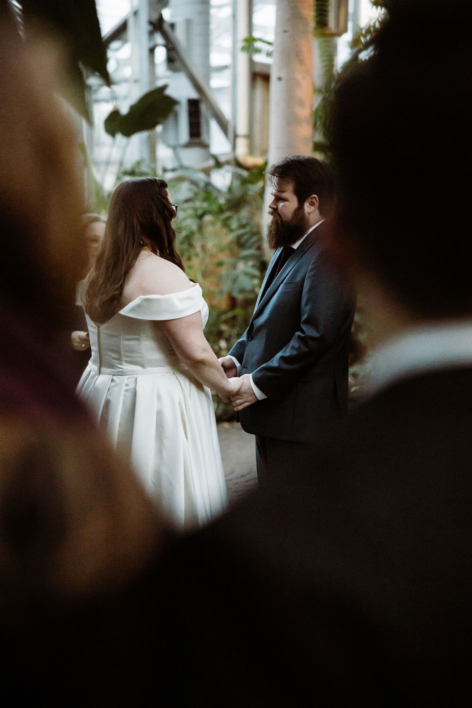 A wedding ceremony moment captured through foliage showing a couple holding hands in an intimate setting.