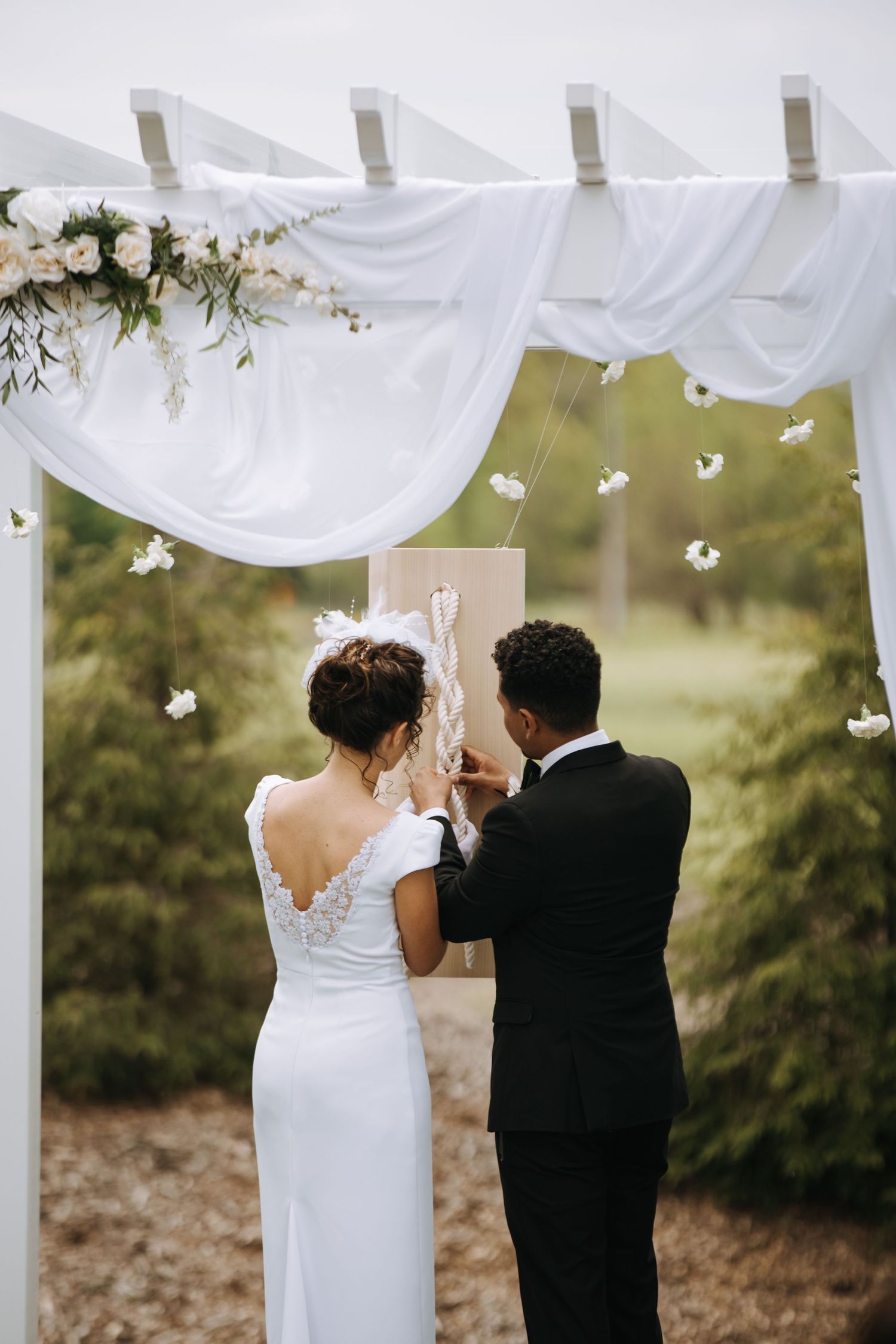 A couple shares a toast under a white draped wedding ceremony arch.