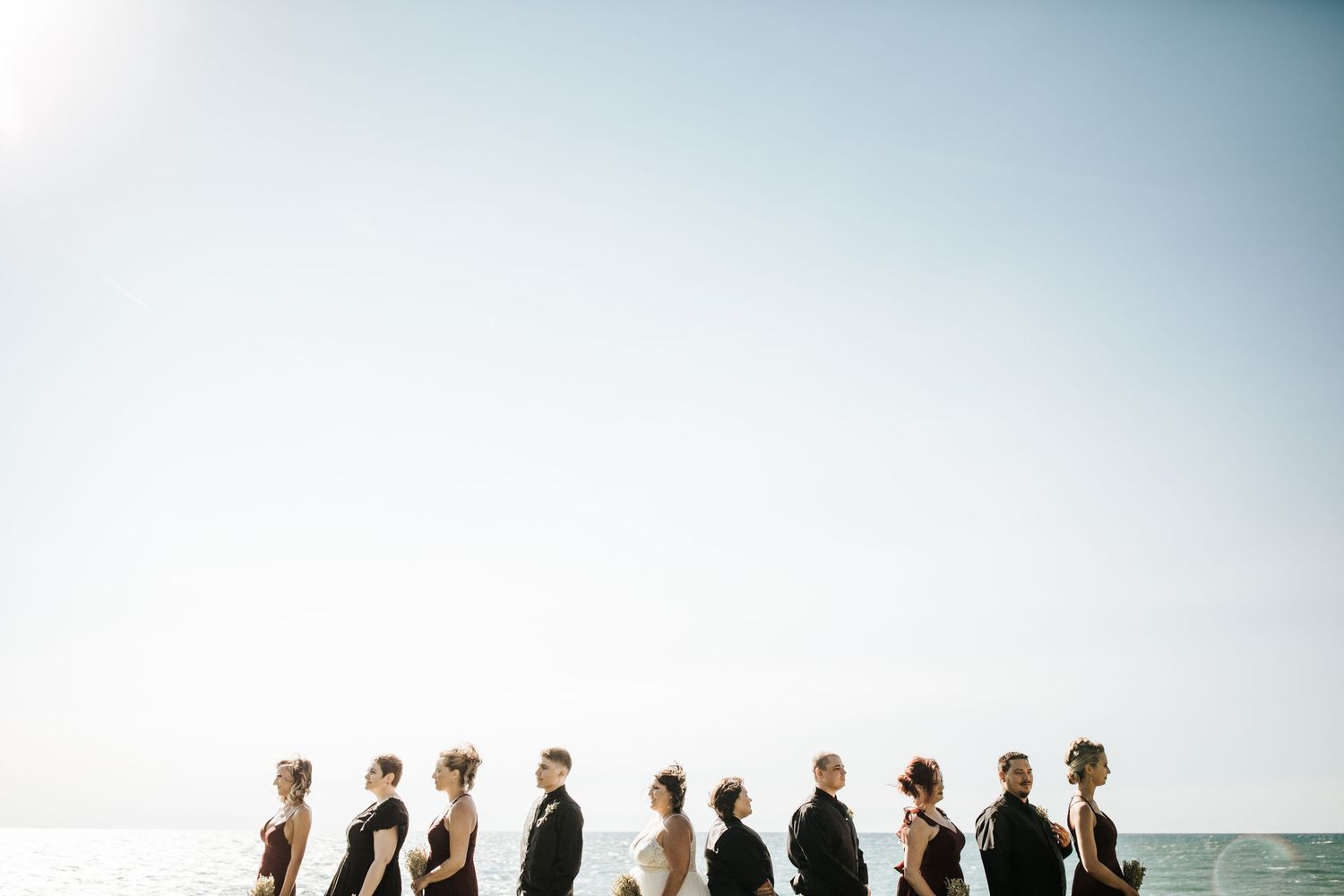 Wedding party stands in silhouette against bright sky and horizon line at beach ceremony.