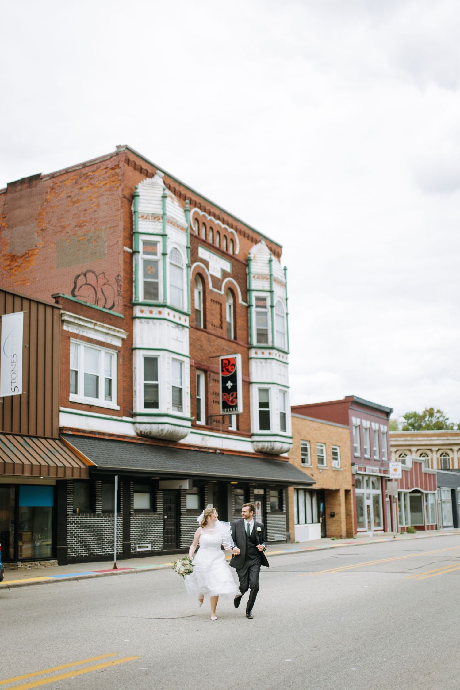 Newlyweds run hand in hand past historic brick buildings in a small town.