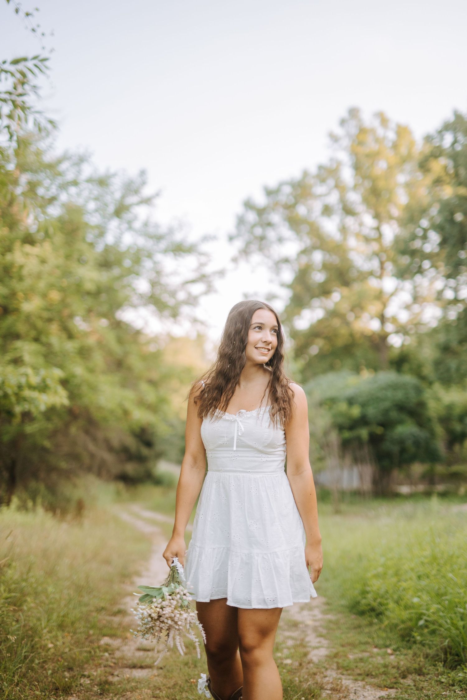 Person in white dress walking through meadow holding wildflowers.