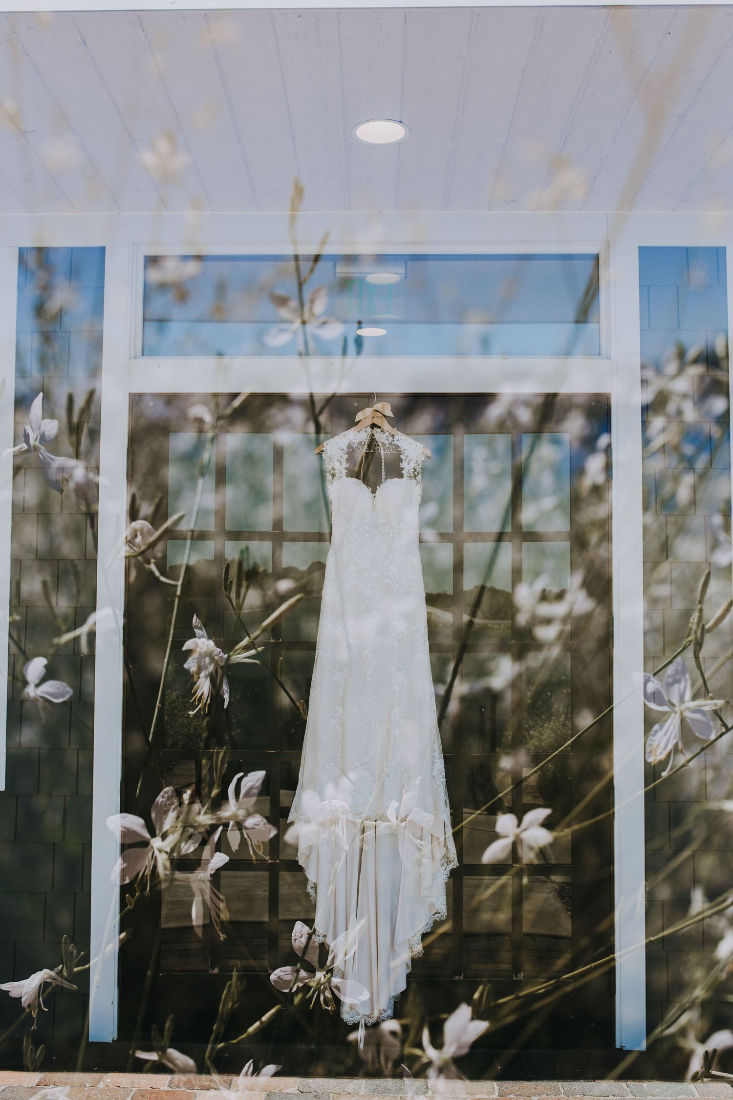 White wedding dress hangs against a window with delicate white flowers blooming in the background.