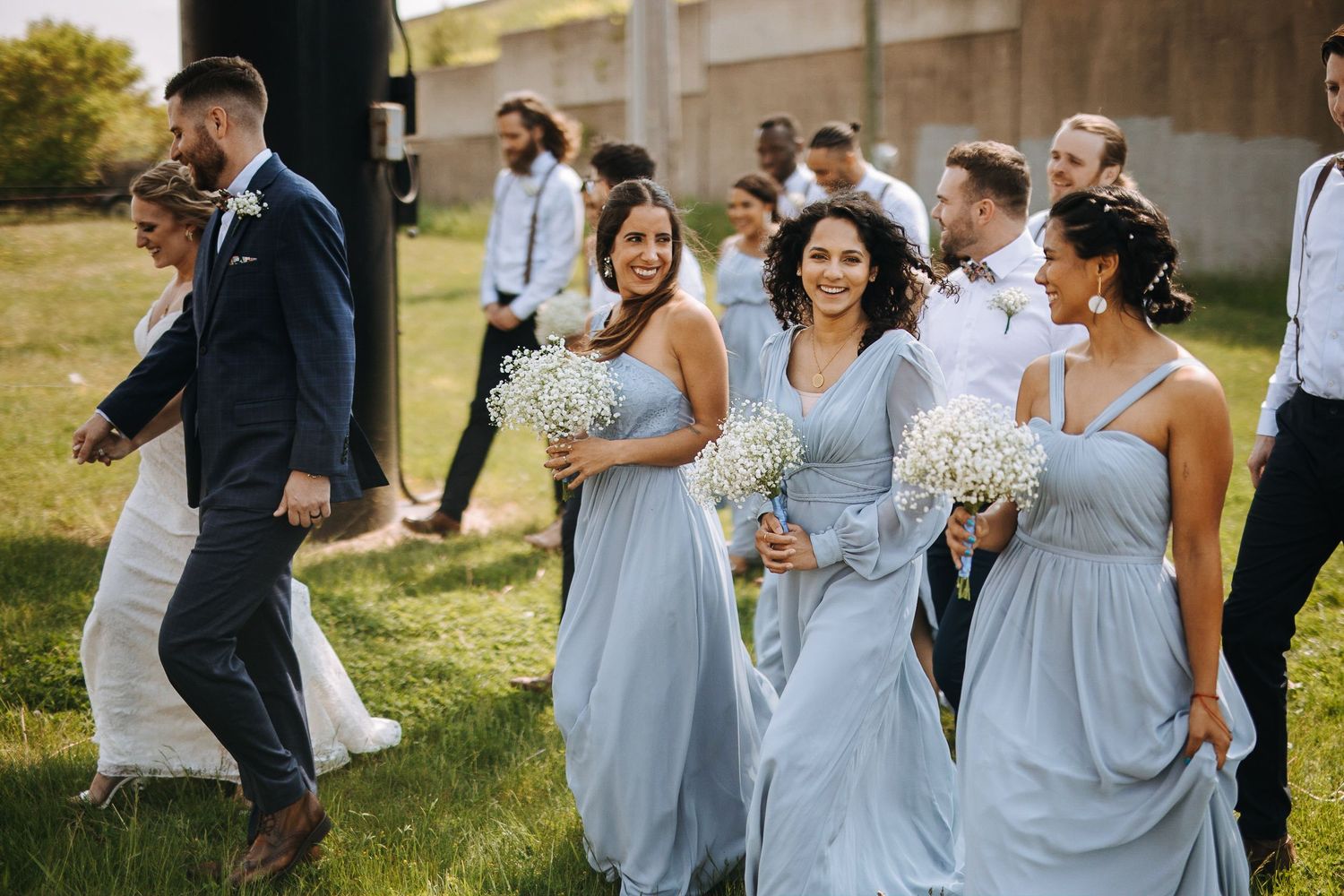 Wedding party poses playfully on grassy lawn in light blue dresses and formal attire.