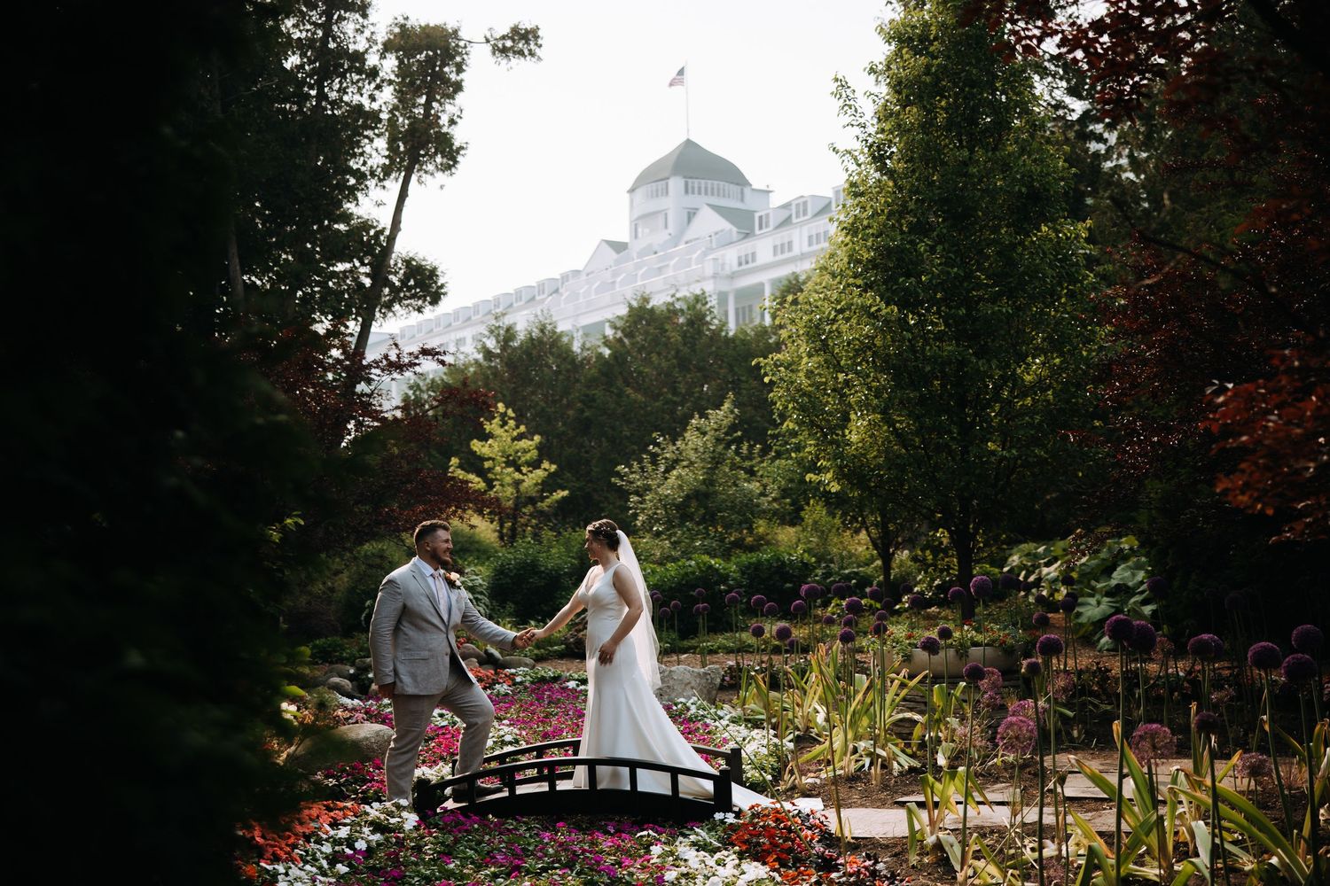 Wedding couple stands on decorative bridge with grand white hotel visible in background.