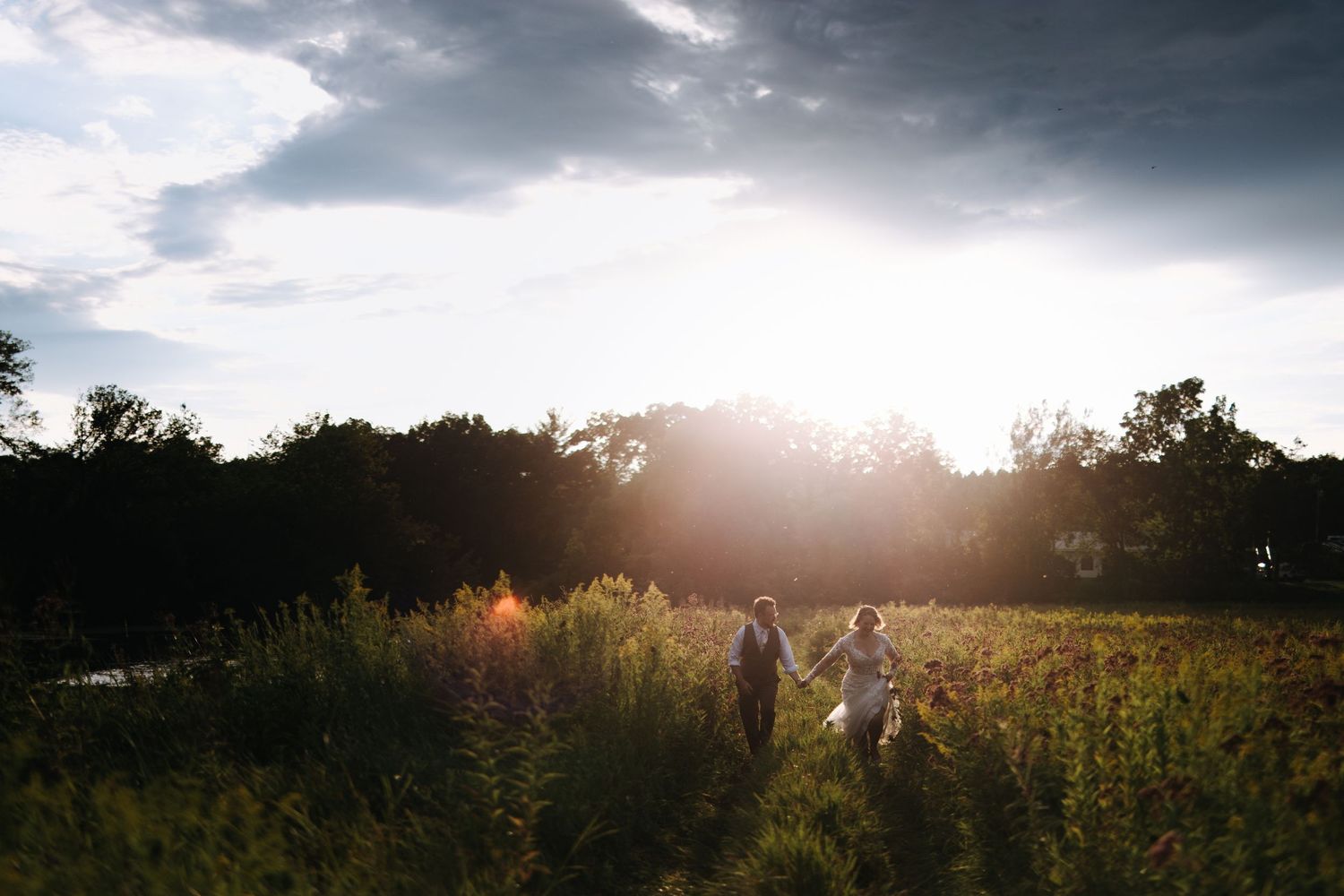 Couple walks through golden field at sunset with dramatic cloudy sky.