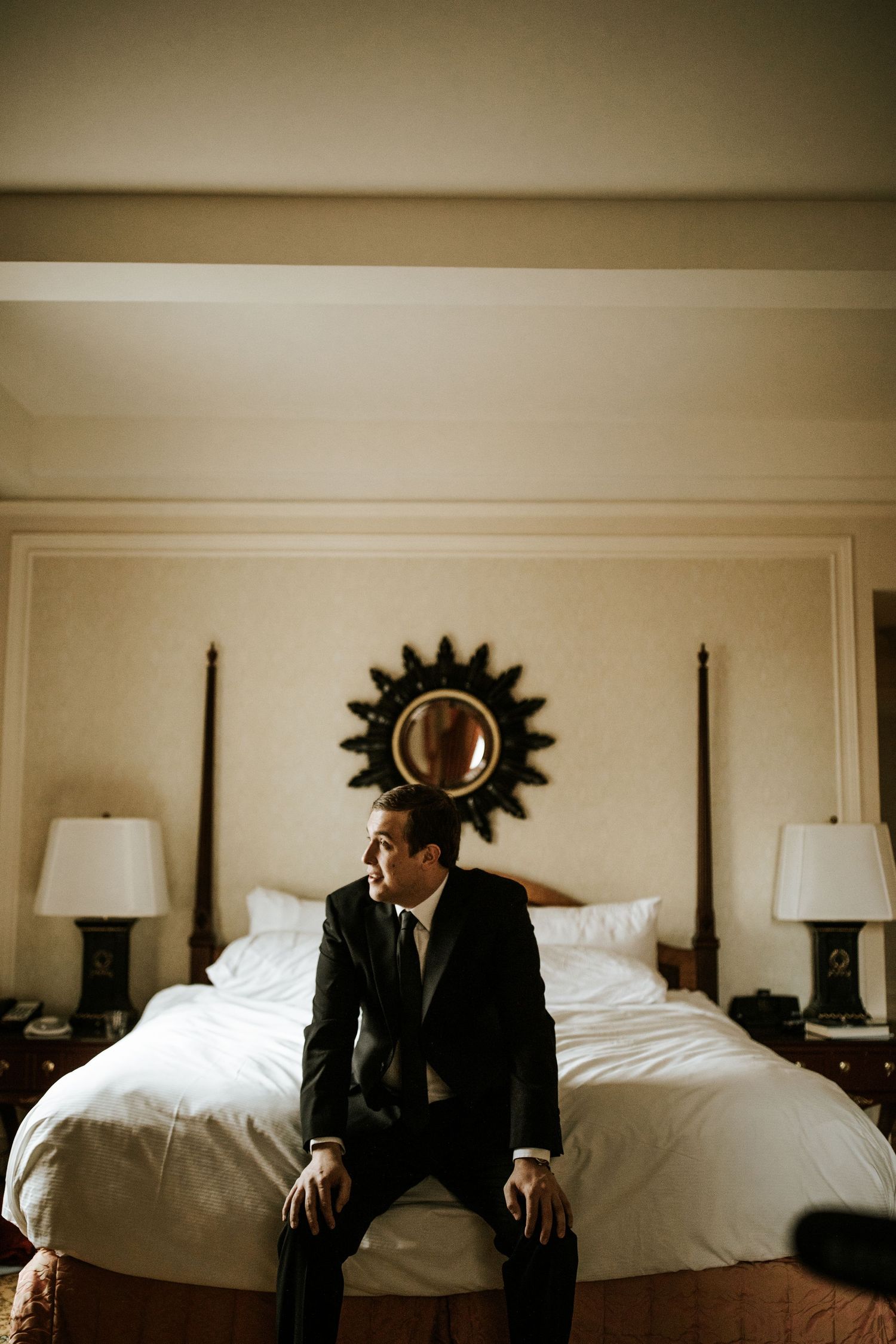 A figure in a dark suit sits contemplatively on the edge of a hotel bed beneath a sunburst mirror.