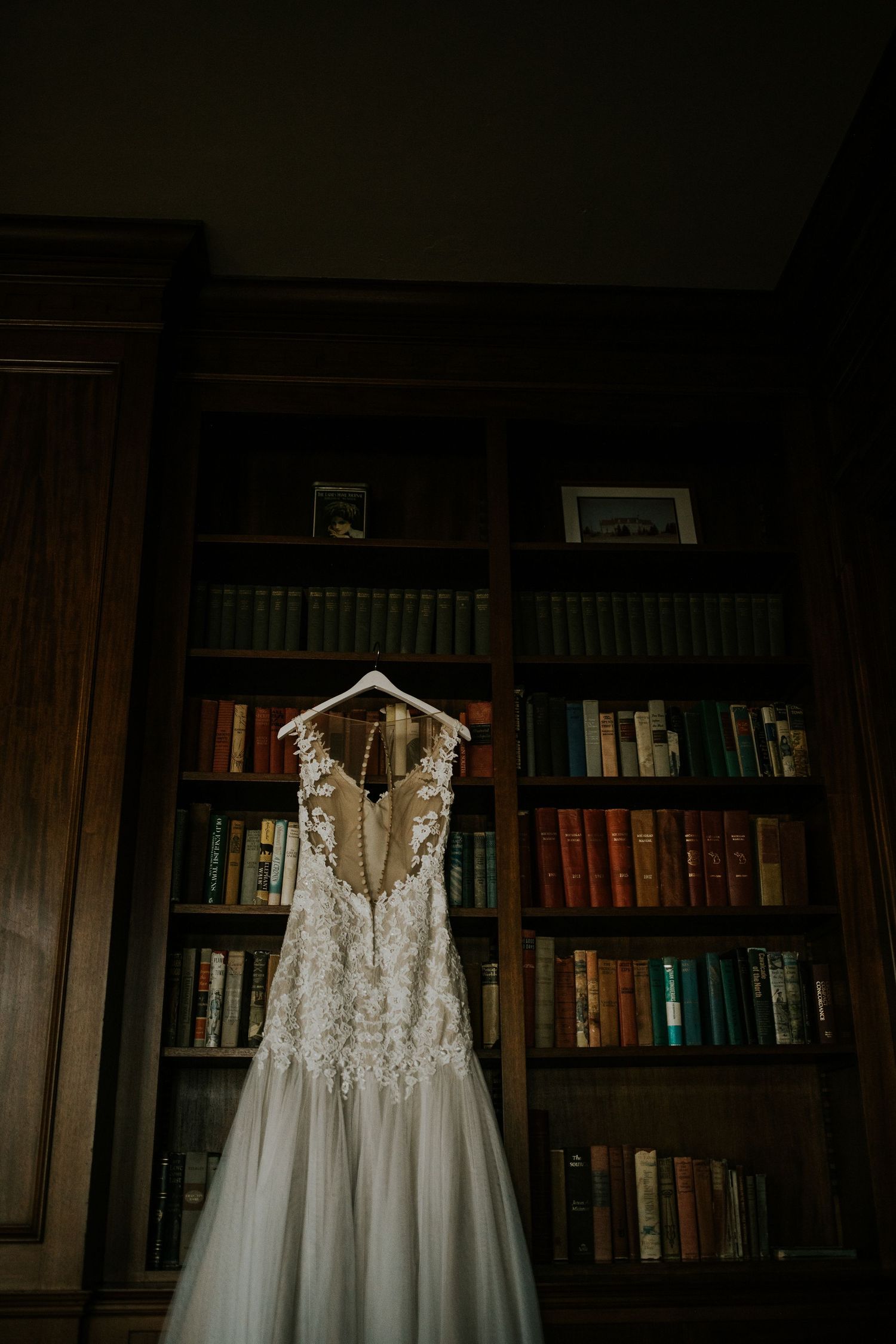 An elegant white lace wedding dress hanging against a backdrop of library bookshelves.