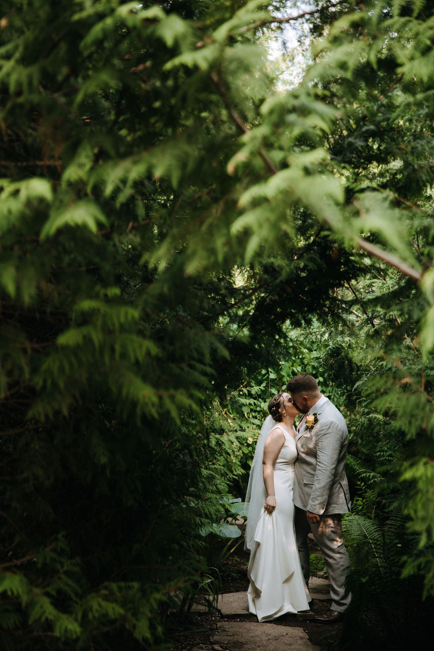 Romantic wedding photo of couple embracing on forest path surrounded by lush greenery.