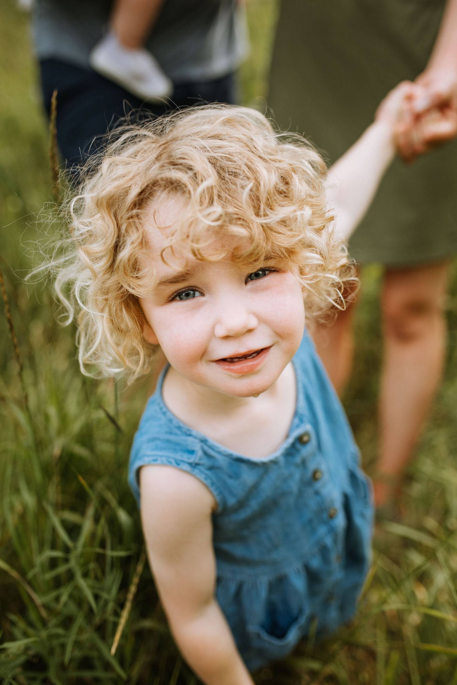 Toddler in blue dress with curly blonde hair smiles brightly in outdoor setting.