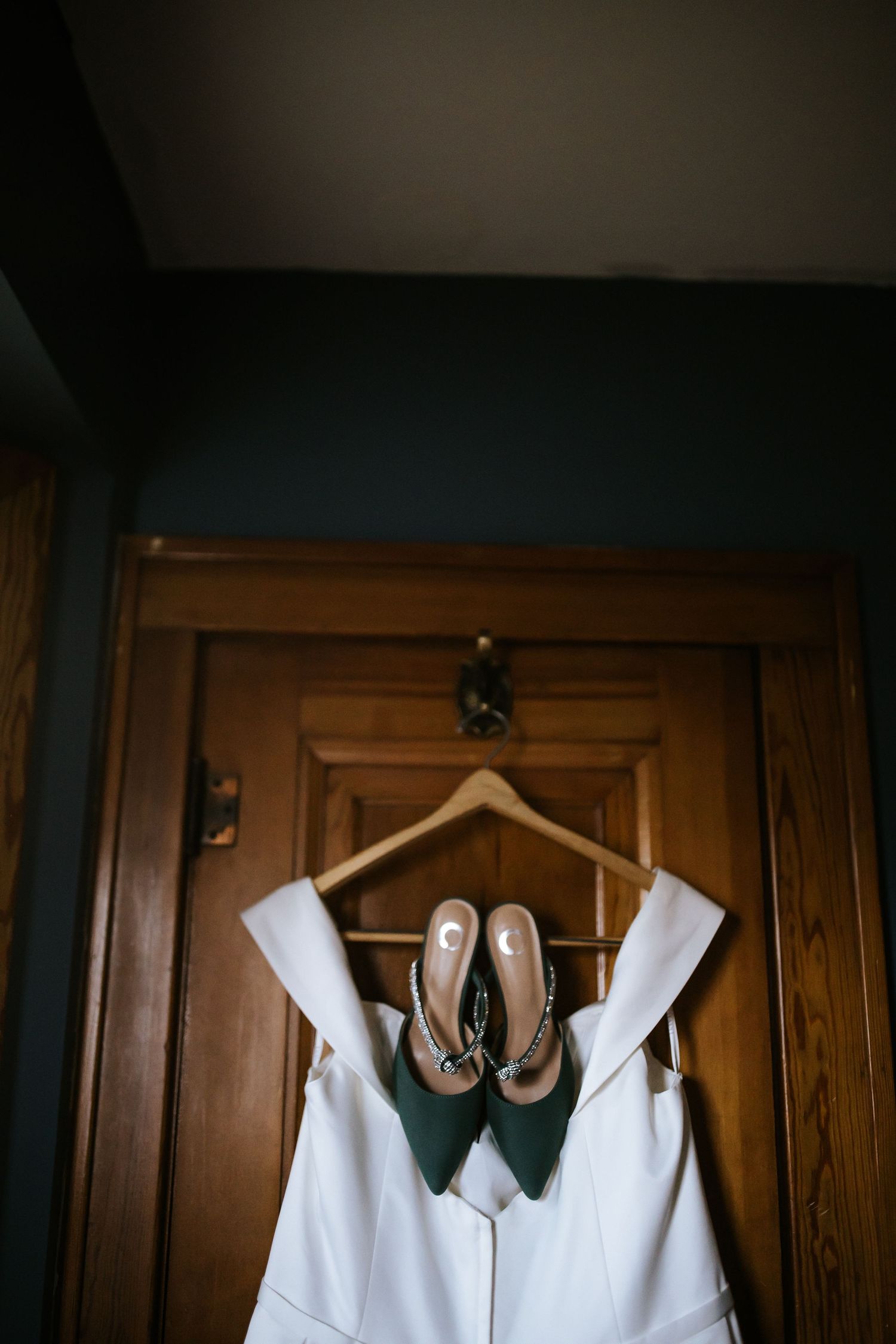 A white dress and green shoes hang on a wooden hanger against a dark wooden door.