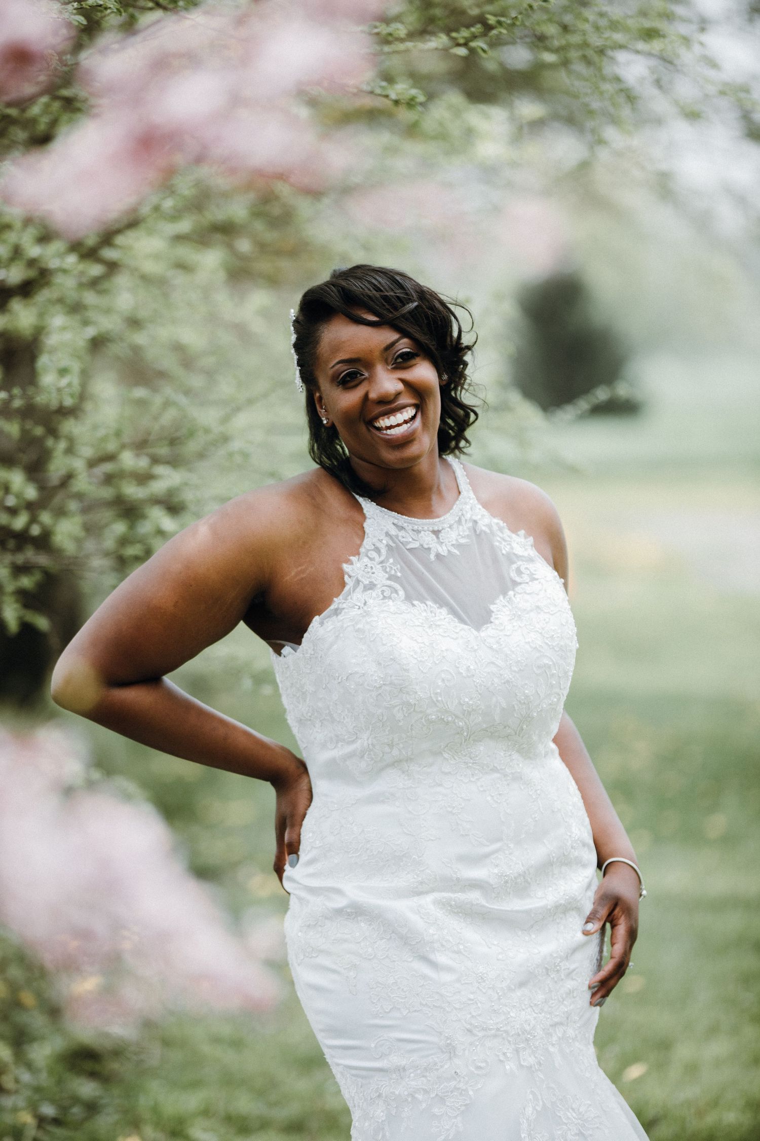 A beautiful bride in a sleeveless lace wedding gown poses joyfully in a garden setting with soft pink blossoms.