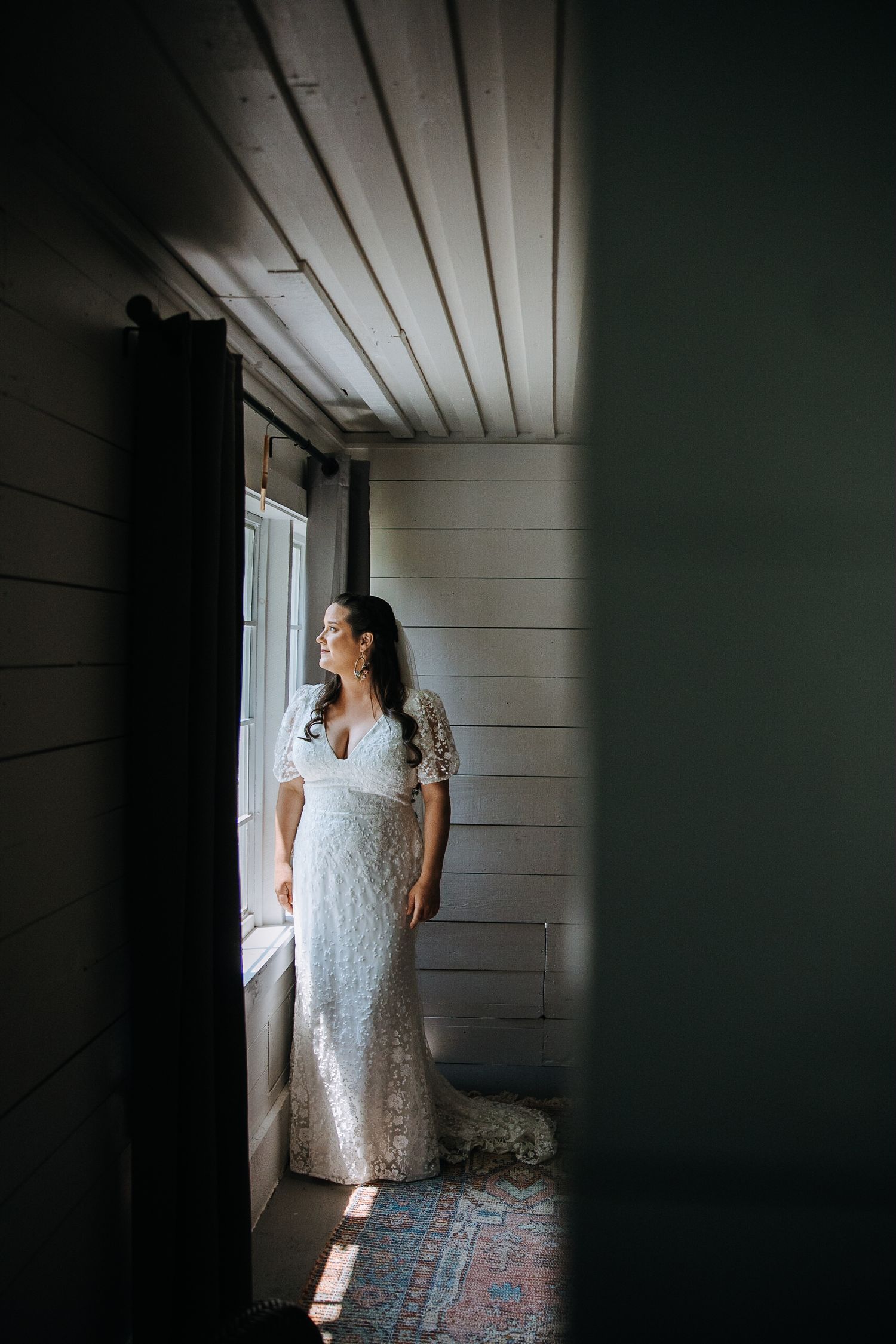Person in white lace dress standing by window in rustic room with dramatic lighting.