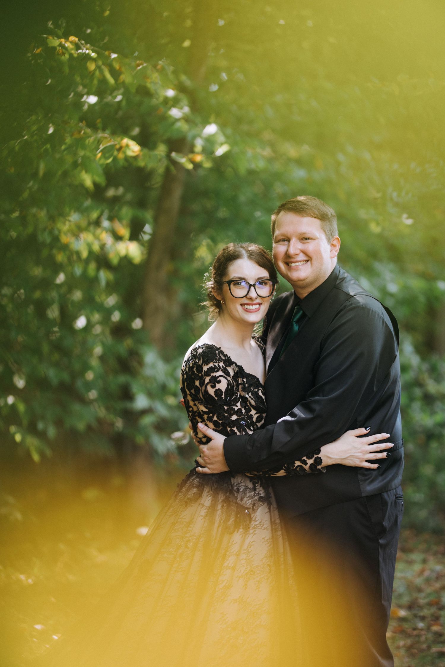 Couple embraces in outdoor setting with golden sunlit foliage in background.