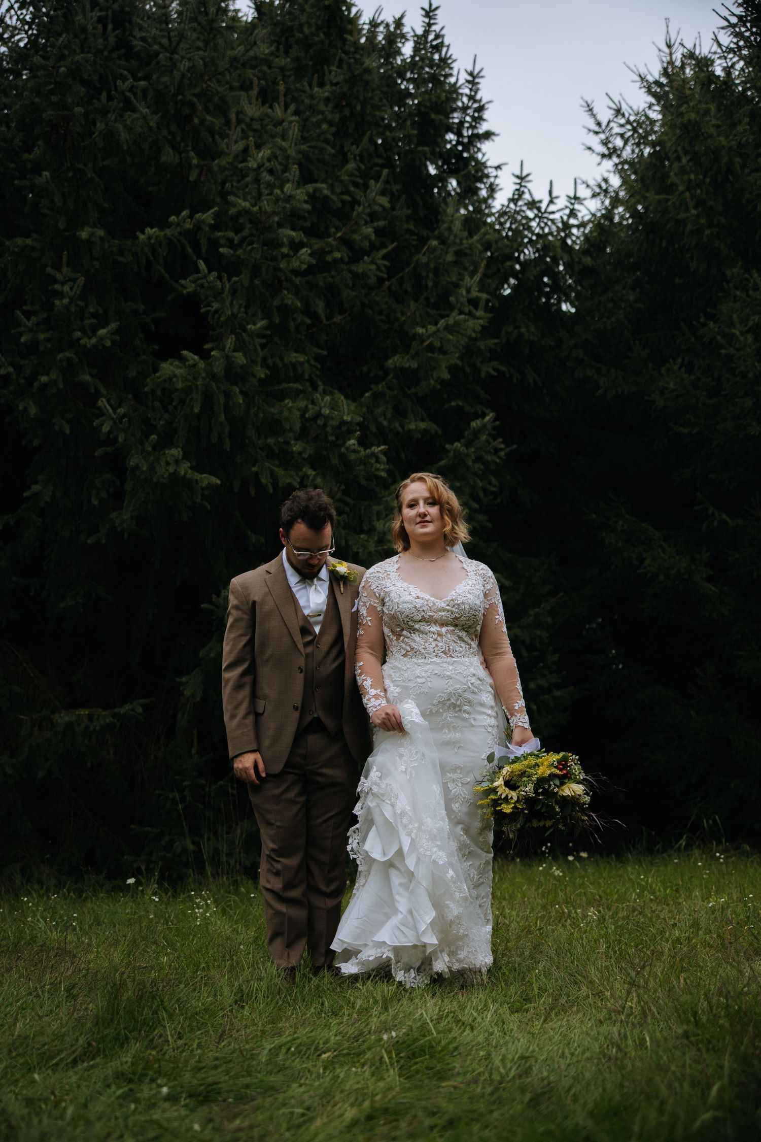 Wedding couple walking through a meadow with evergreen trees in the background.